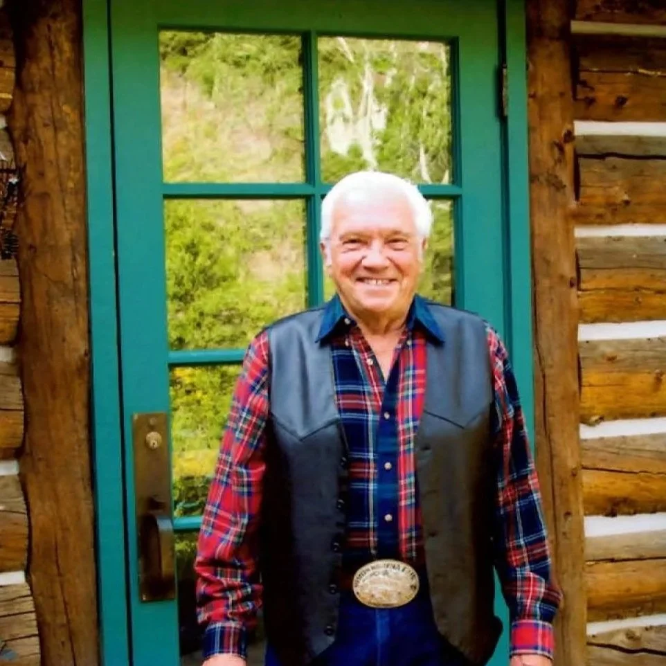A smiling elderly man with white hair wearing a plaid shirt, leather vest, and belt buckle standing in front of a green-framed window on a rustic wooden cabin exterior.