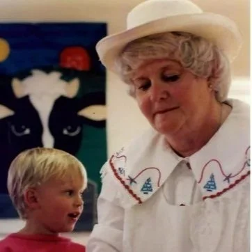 An elderly woman with white hair wearing a white hat and dress, talking to a young boy with blonde hair wearing a red shirt, in front of a colorful painting.