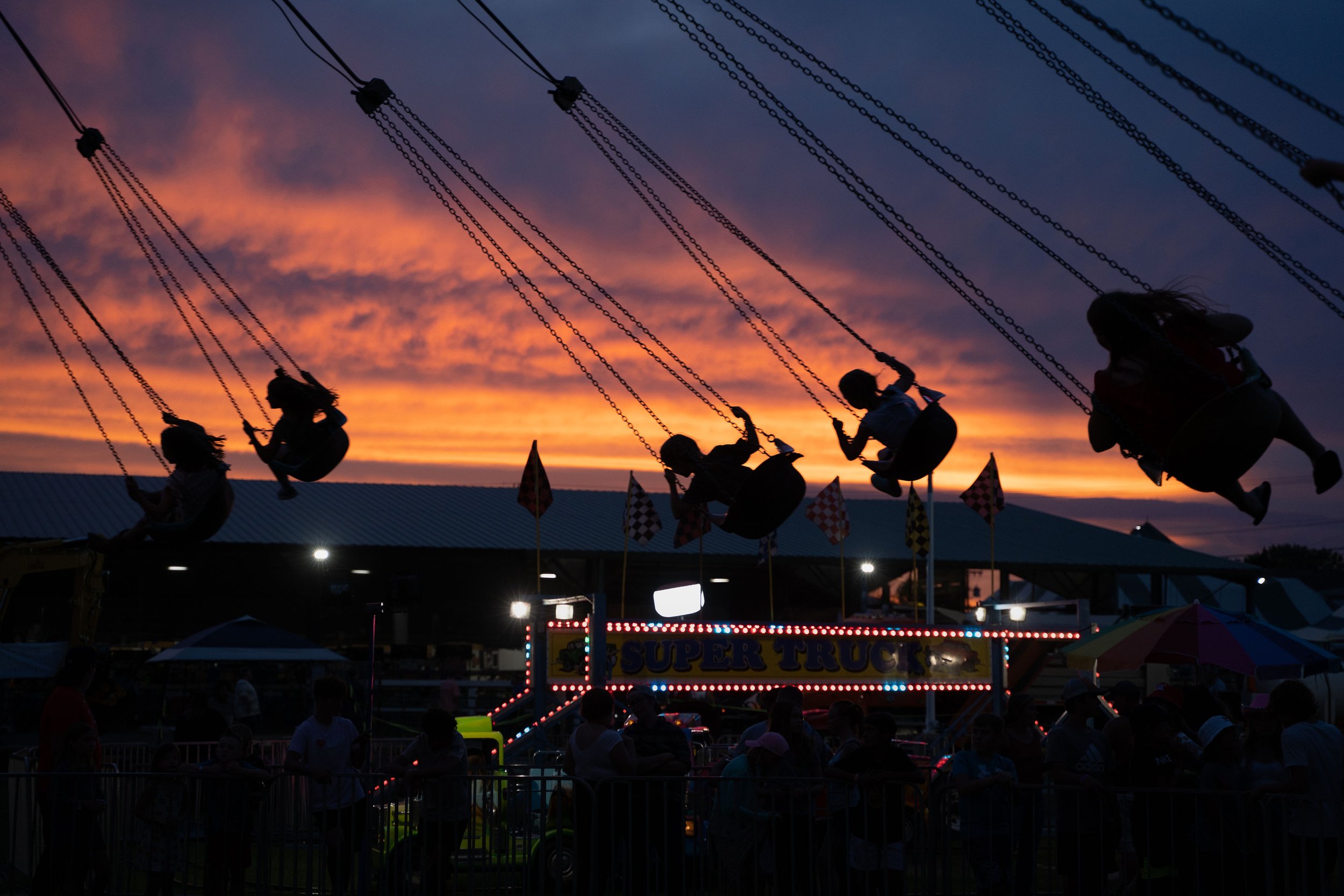 Children riding a swing amusement ride at dusk with a colorful sunset sky and fairground stalls in the background.