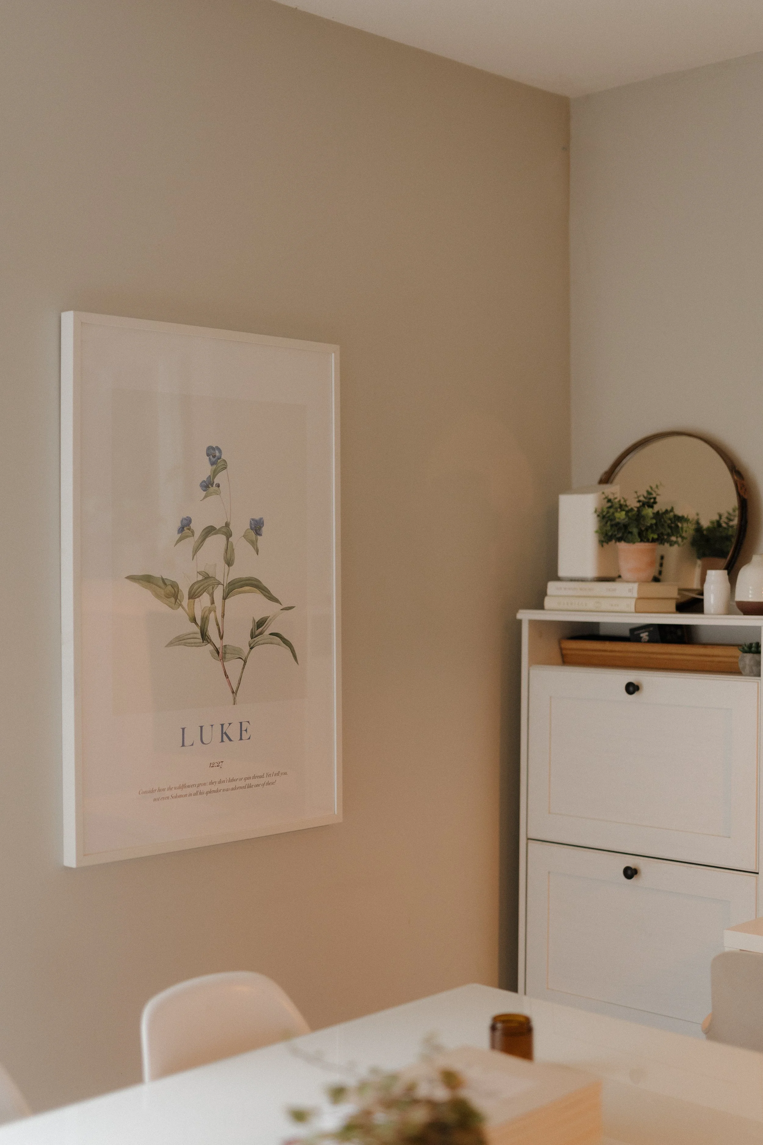 A room with beige and white walls, featuring a framed botanical print of a plant named Luke, a white dresser with a potted plant, books, and decorative items, and a round mirror. Part of a table and chair are visible in the foreground.