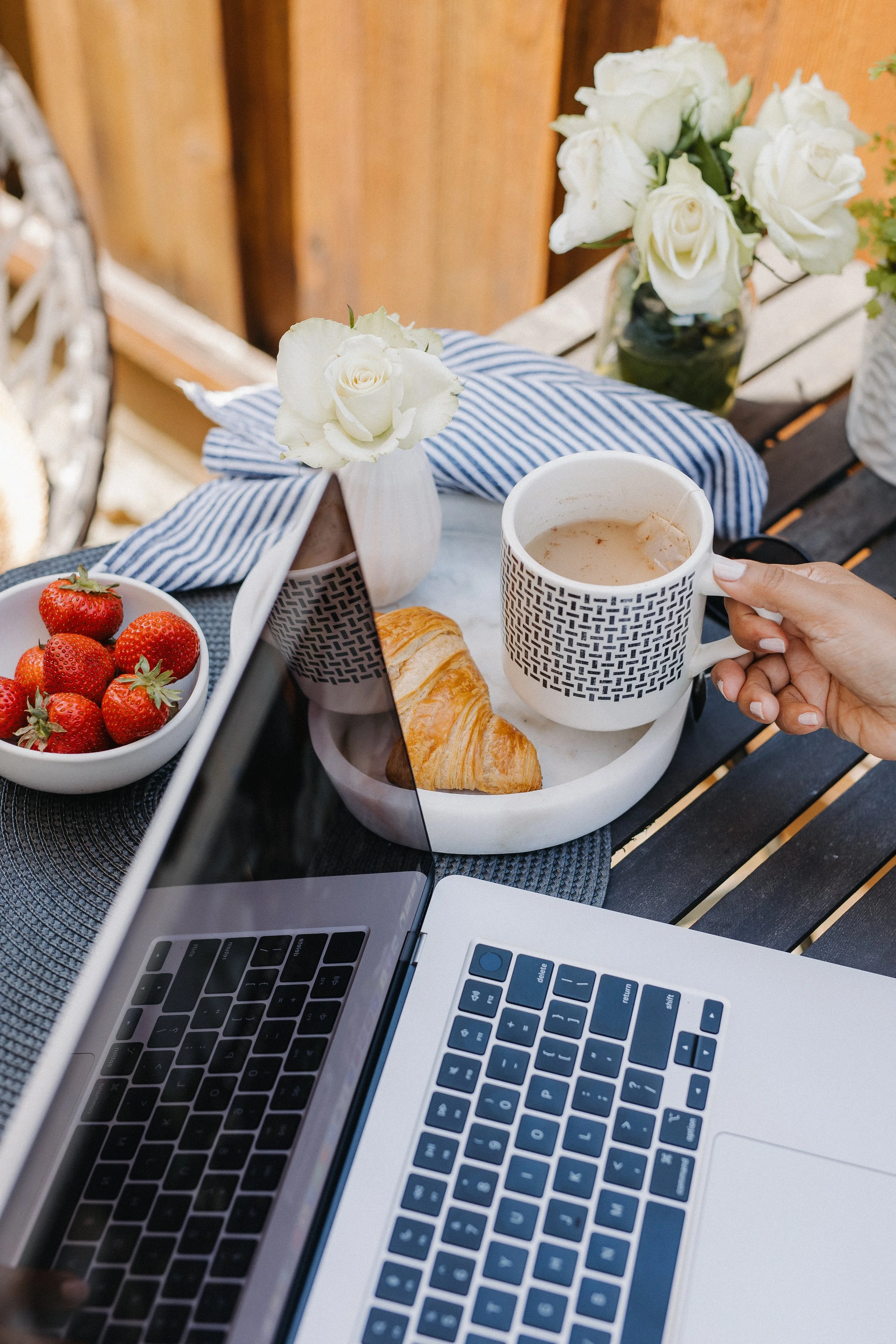 A breakfast tray on a table with a laptop, a cup of coffee, fresh strawberries, a croissant, and white roses in a vase, set outdoors with a wooden fence in the background.