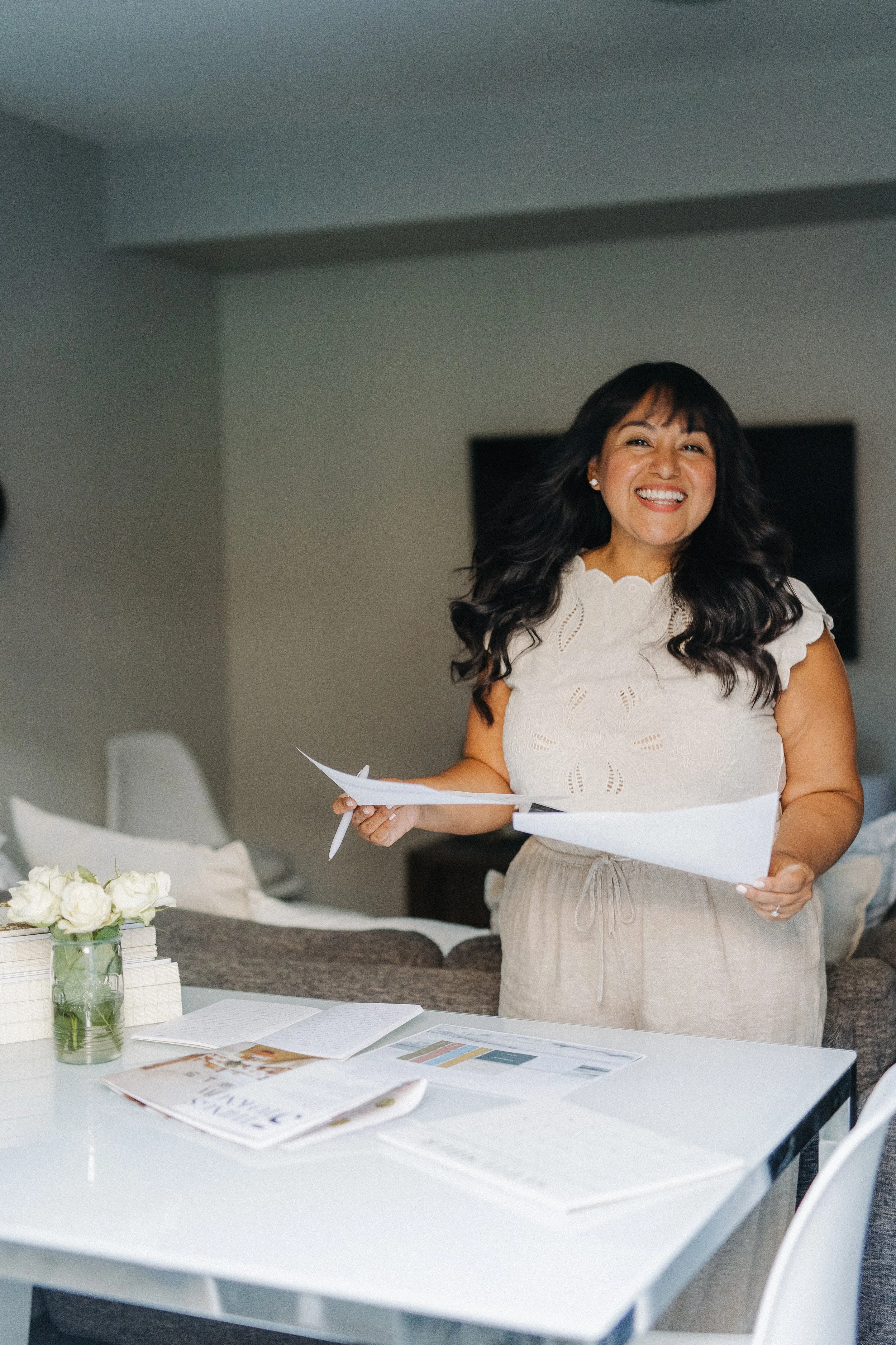 A woman with dark hair and a white eyelet blouse is smiling while holding papers and standing at a table with documents, a vase of white roses, and a colorful chart.