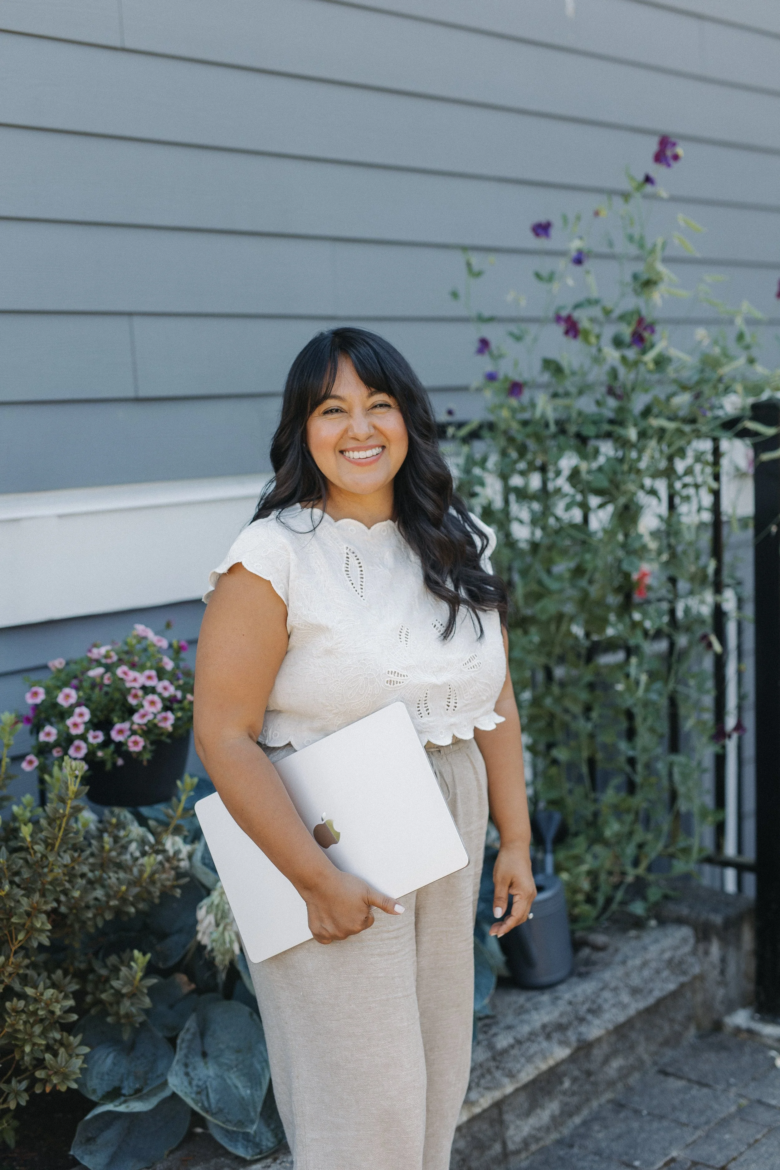 A woman with long dark hair, wearing a white embroidered blouse and beige pants, is holding a closed silver MacBook laptop and smiling in front of a house with grey siding and flowering plants.