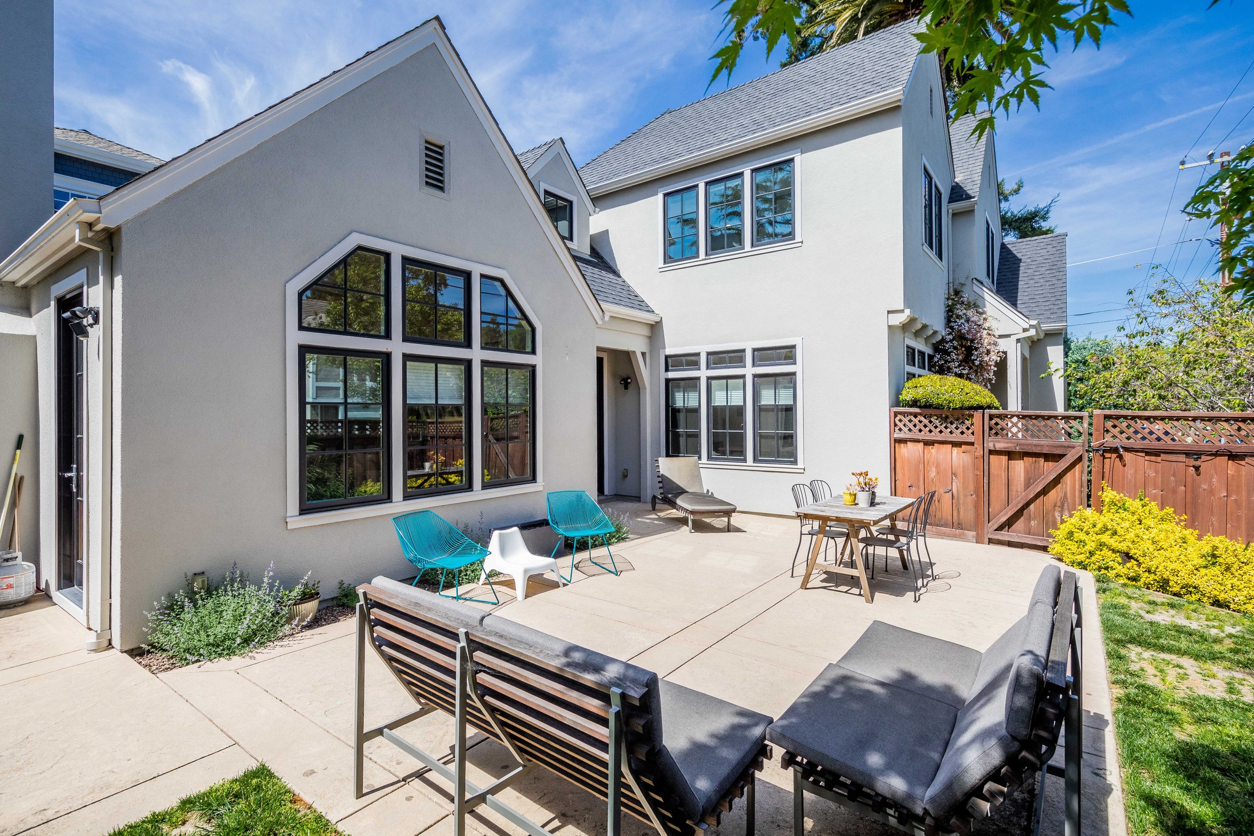 A backyard patio with outdoor furniture, including a sofa, chairs, and a table, surrounded by a wooden fence and neighboring houses under a clear blue sky.