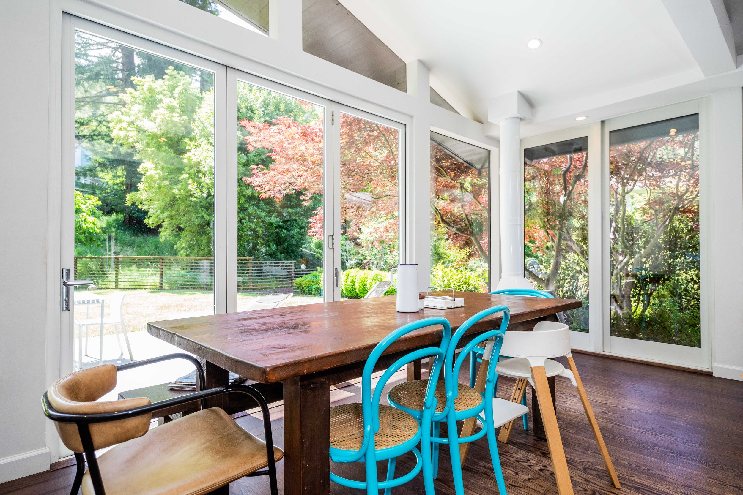 Bright dining area with large windows showing green and red foliage outside, a wooden table surrounded by chairs in different styles and colors, and a high white chair.