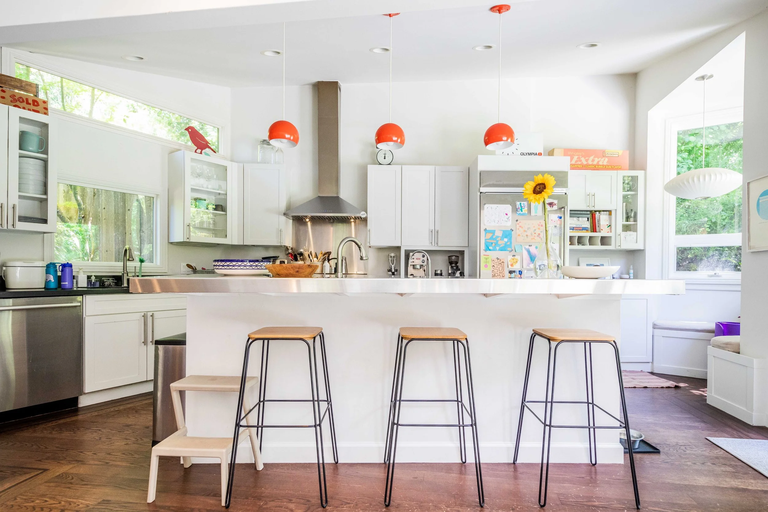 Bright kitchen with white cabinets, stainless steel appliances, and wooden barstools at a white island. Red pendant lights hang over the island. Large windows bring in natural light.