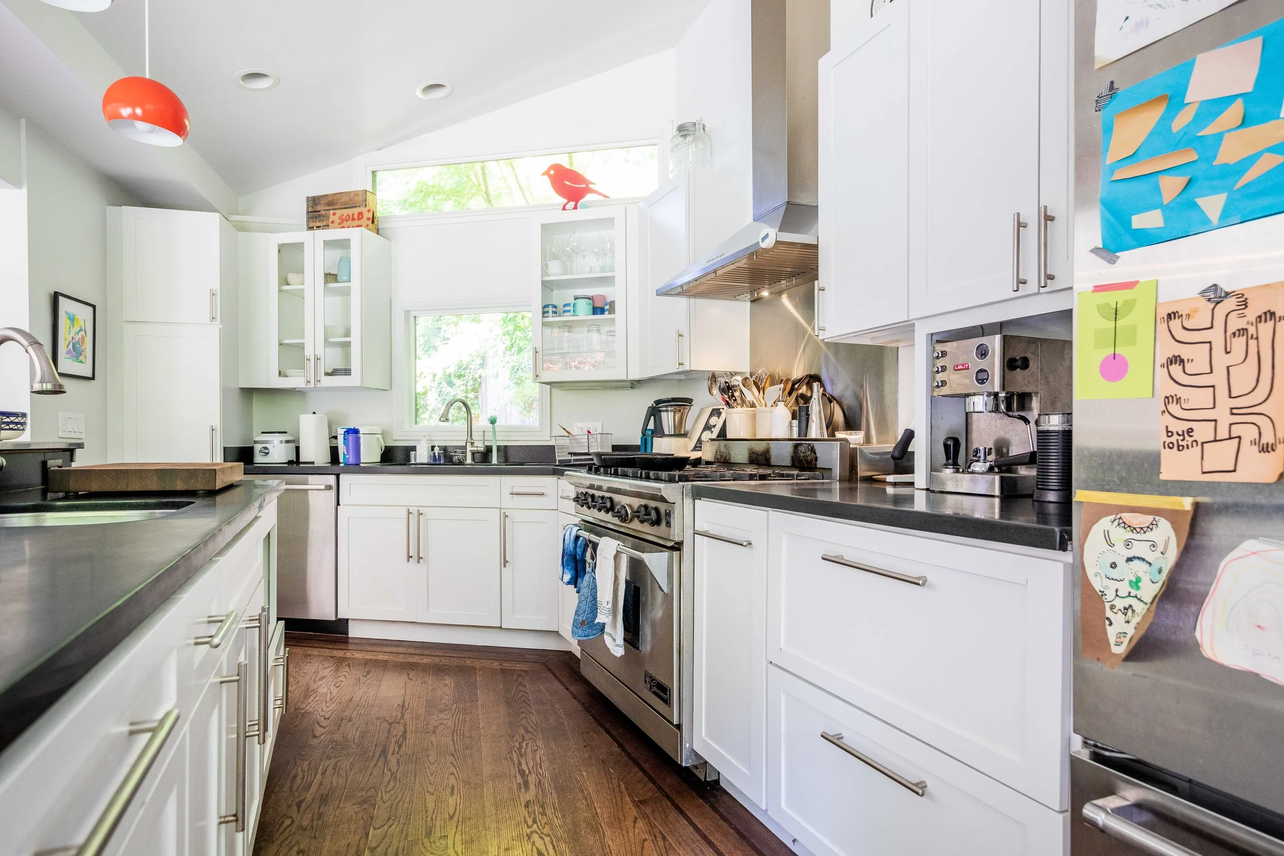 Modern white kitchen with black countertops, stainless steel appliances, and hardwood flooring. There are colorful children's drawings on the refrigerator and a small red bird figurine on a high shelf near the window.