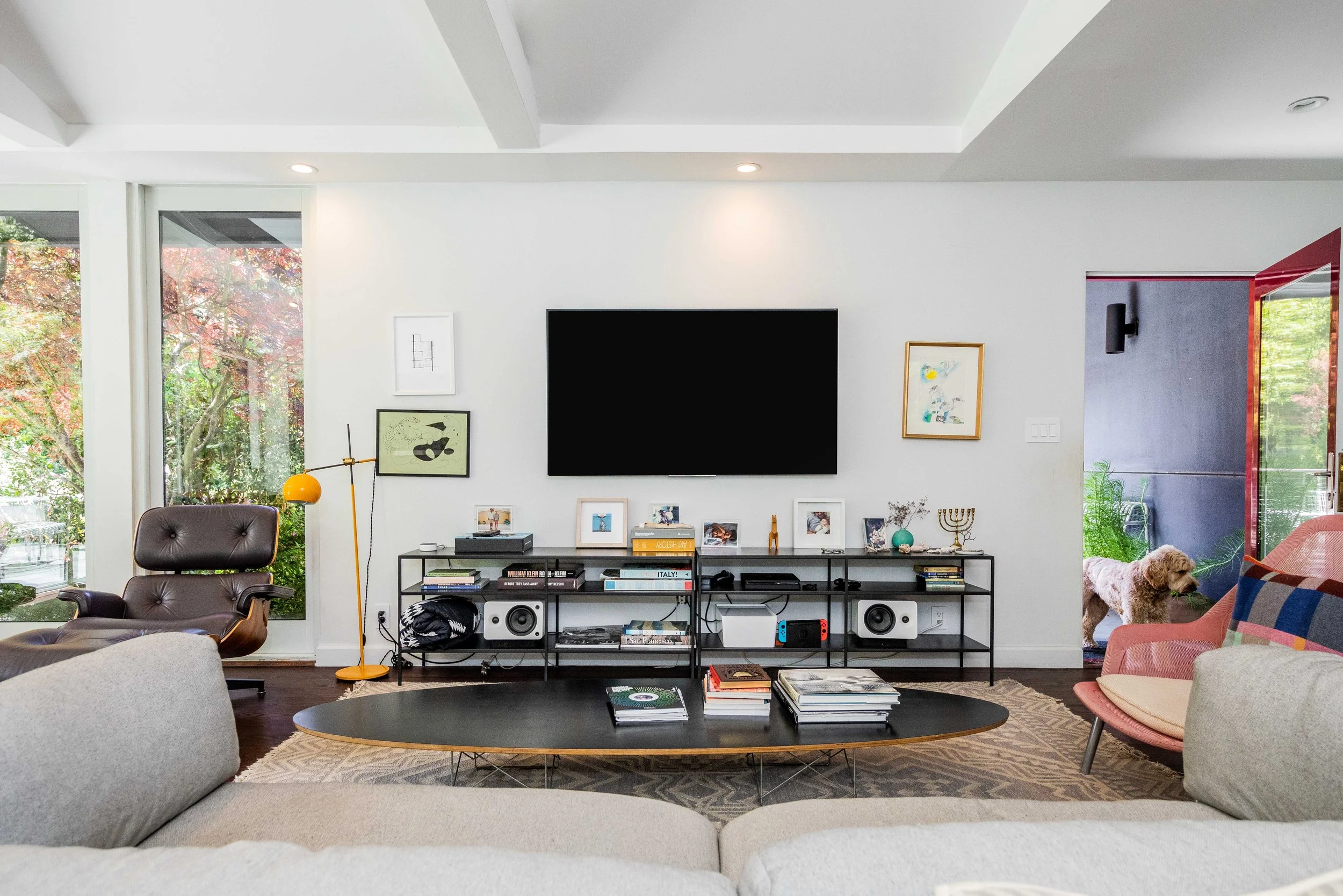 Living room with modern decor, large window showing trees outside, TV on wall, black metal console with books, pictures, and speakers, pink armchair with dog, and white sofa.