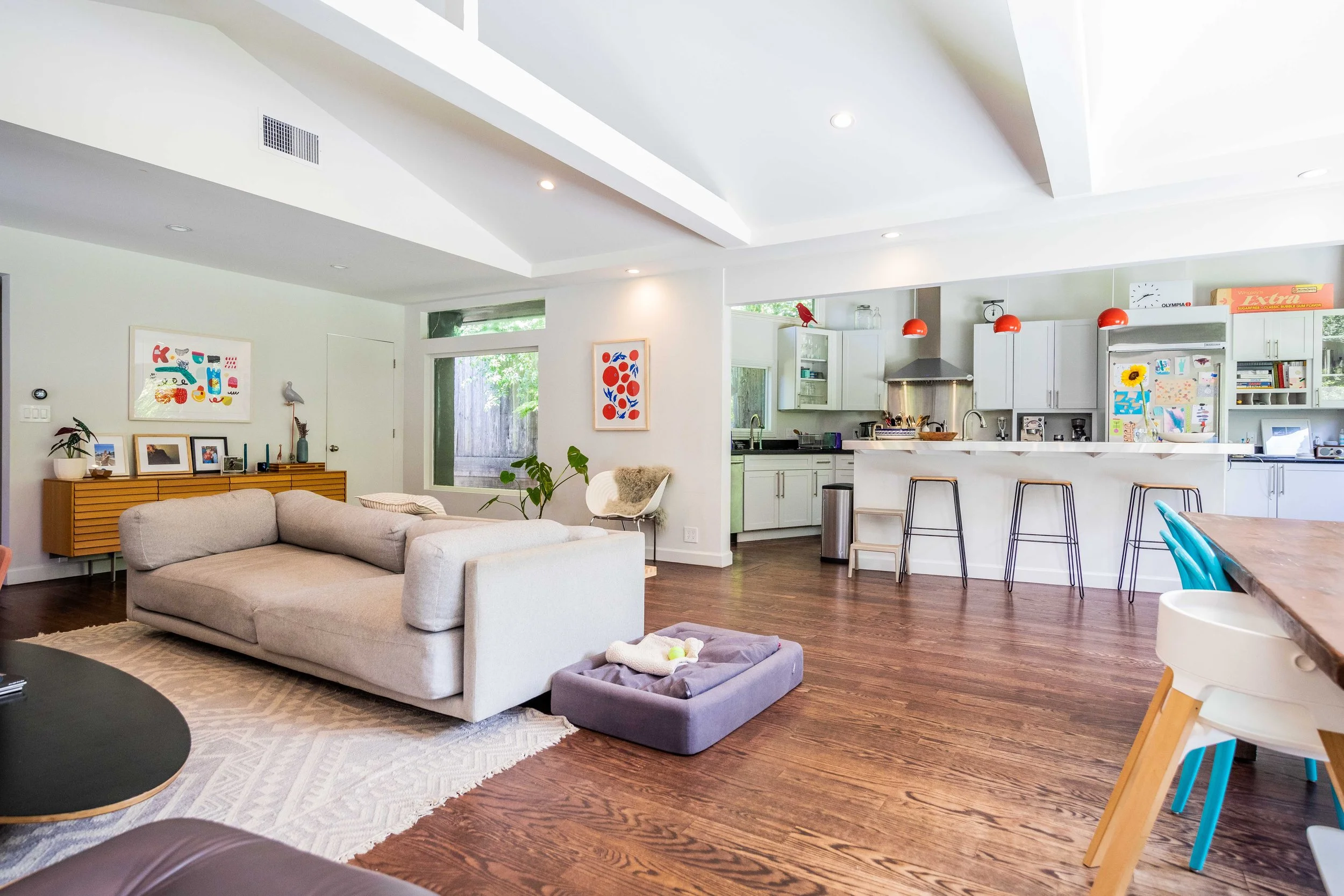 Open-concept living room and kitchen in a modern home with white walls, hardwood floors, a beige sectional sofa, colorful artwork, and brightly colored chairs at the dining table.