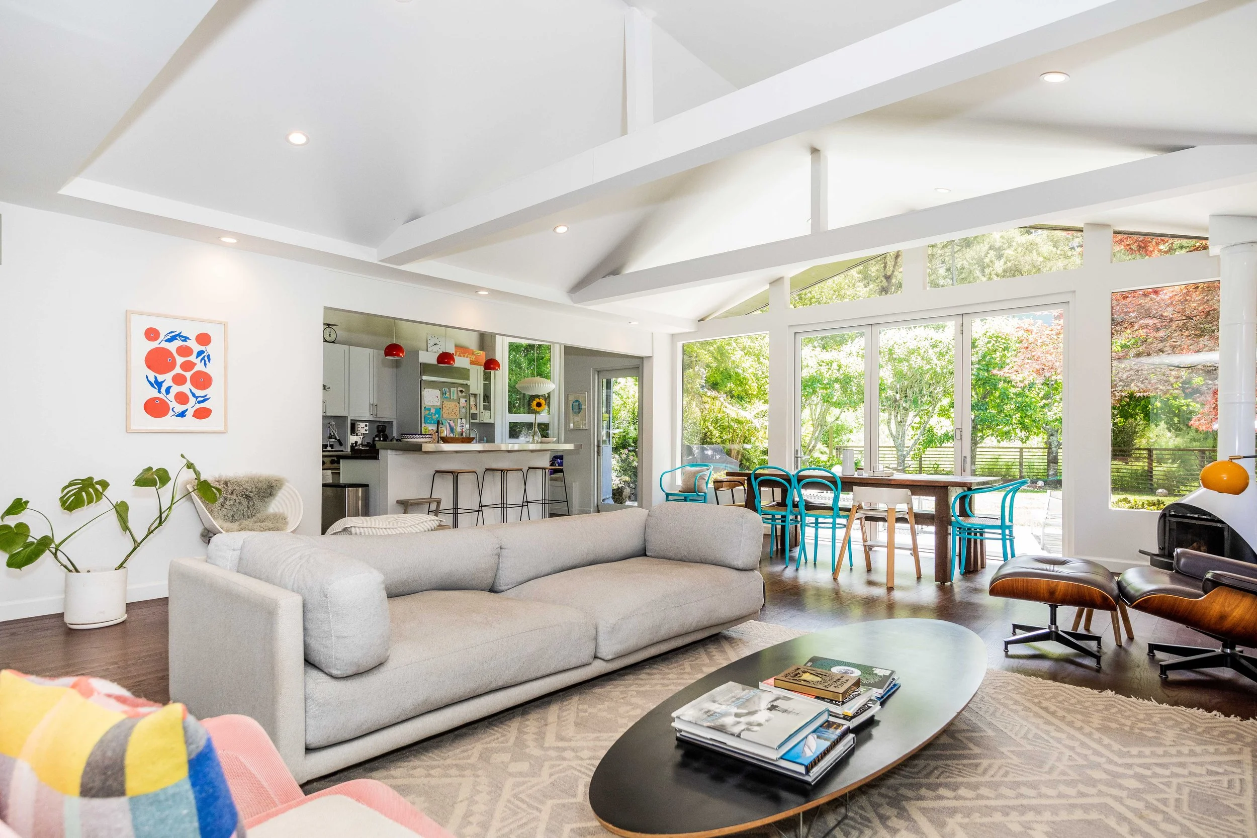 Bright living room with white walls and large windows overlooking lush green trees, featuring a beige sofa, a black coffee table with magazines, a colorful striped chair, and a modern lounge chair with an ottoman, with a view of a dining area and kit