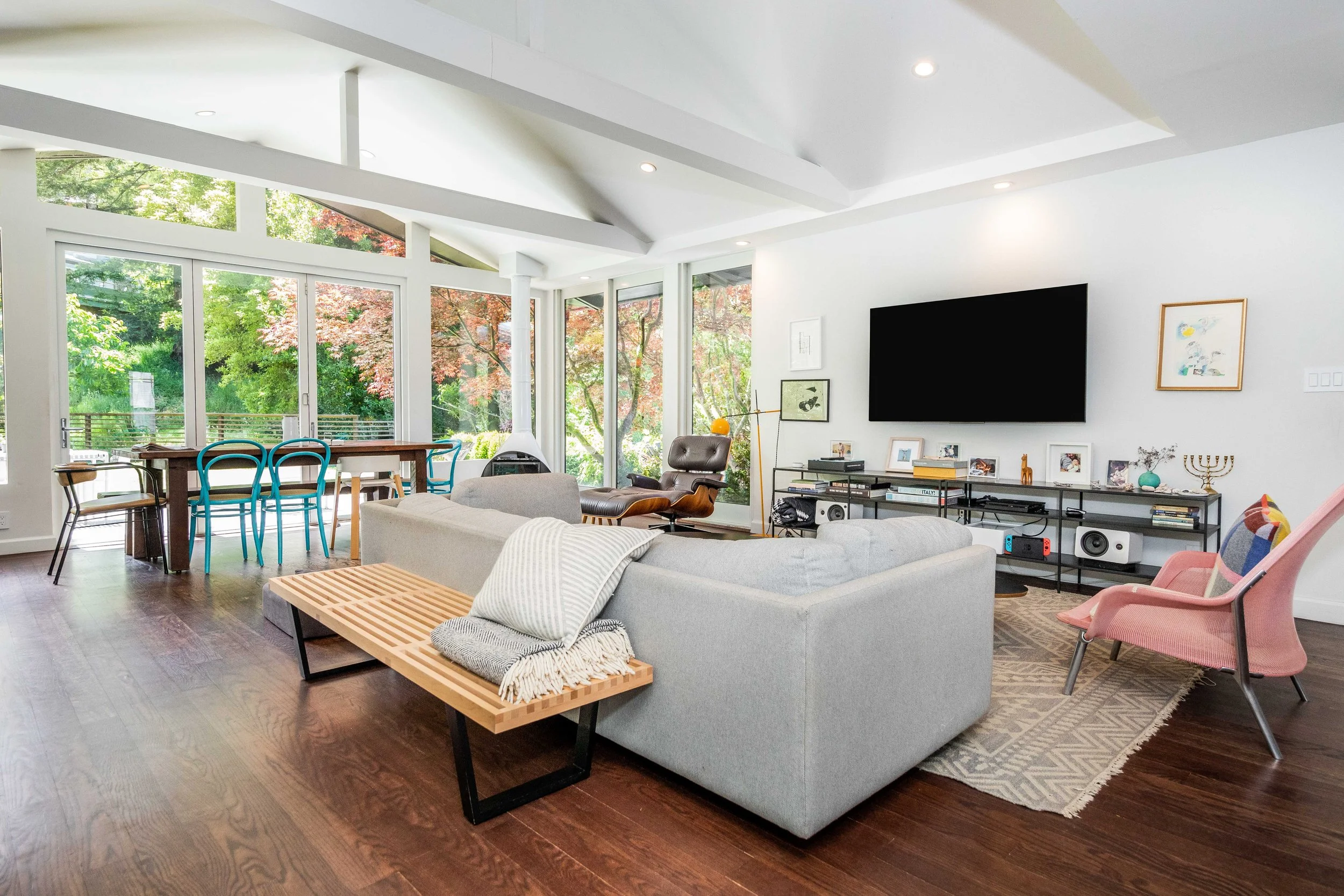 Modern living room with large glass windows, gray sectional sofa, pink armchair, wooden dining table with blue chairs, wall-mounted TV, and decorative items.