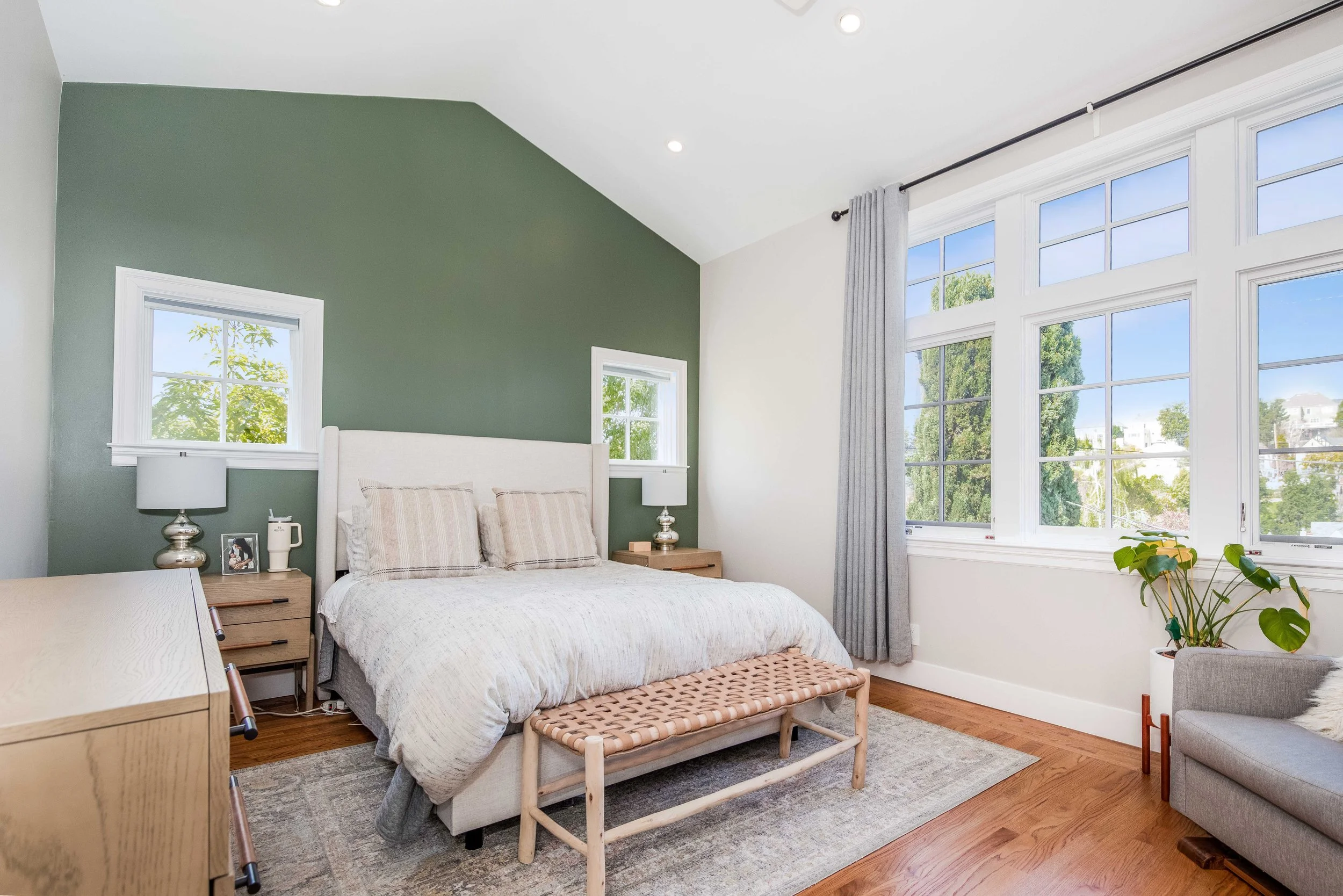 A bedroom with a green accent wall, a large window with white framing and gray curtains, a bed with white and beige bedding, two bedside tables with lamps, a bench at the foot of the bed, and a gray armchair with a potted plant nearby.