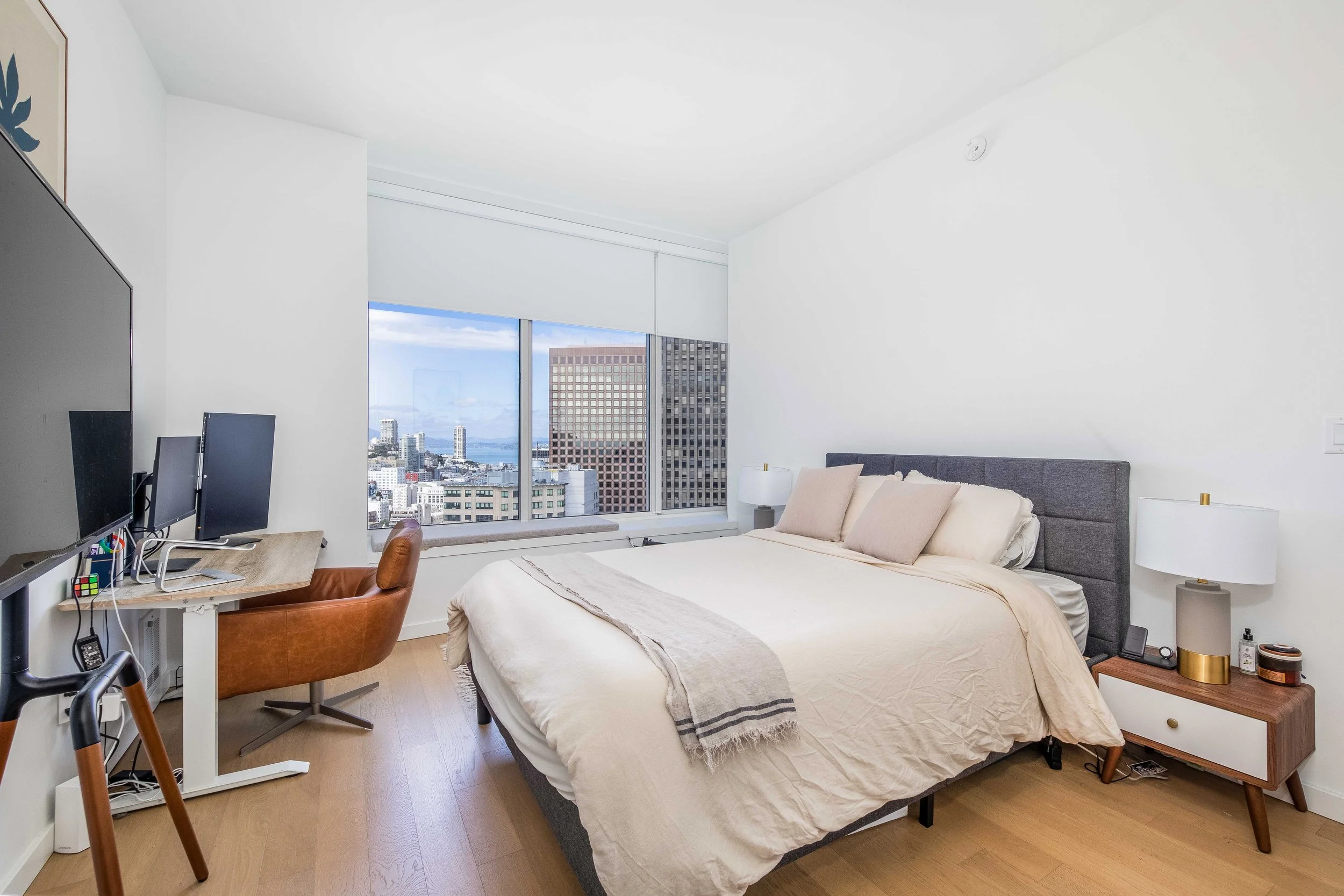 A modern bedroom with a large window showing a city skyline, a bed with beige and cream bedding, a nightstand with a white lamp and personal items, and a workspace with a computer monitor and brown leather chair.