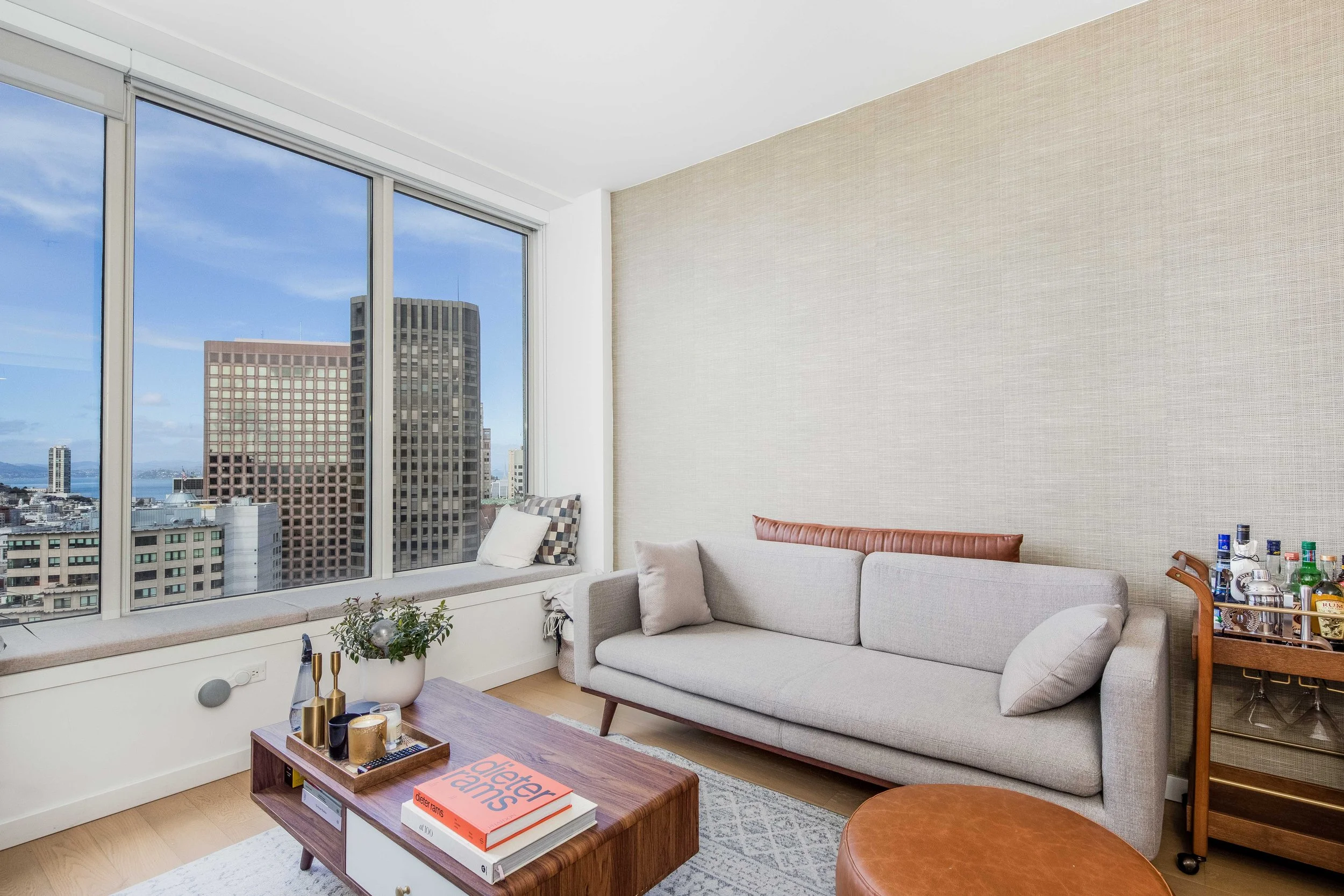Living room with a large window showing city buildings, a light gray sofa with cushions, a wooden coffee table with books, a small bar cart with bottles, and decorative items.