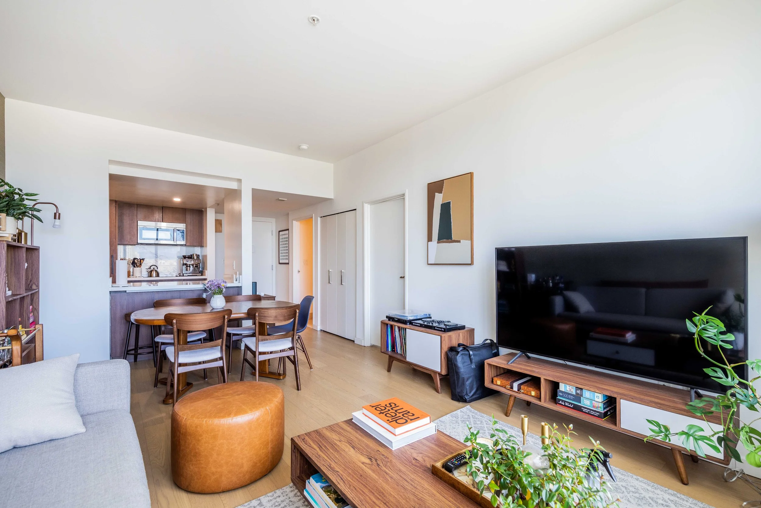 Modern living room with a large flat-screen TV, wooden media console, potted plants, a coffee table with books, a light gray sofa with pillows, and a round leather ottoman. In the background, there's a dining area with a wooden table and chairs, and 