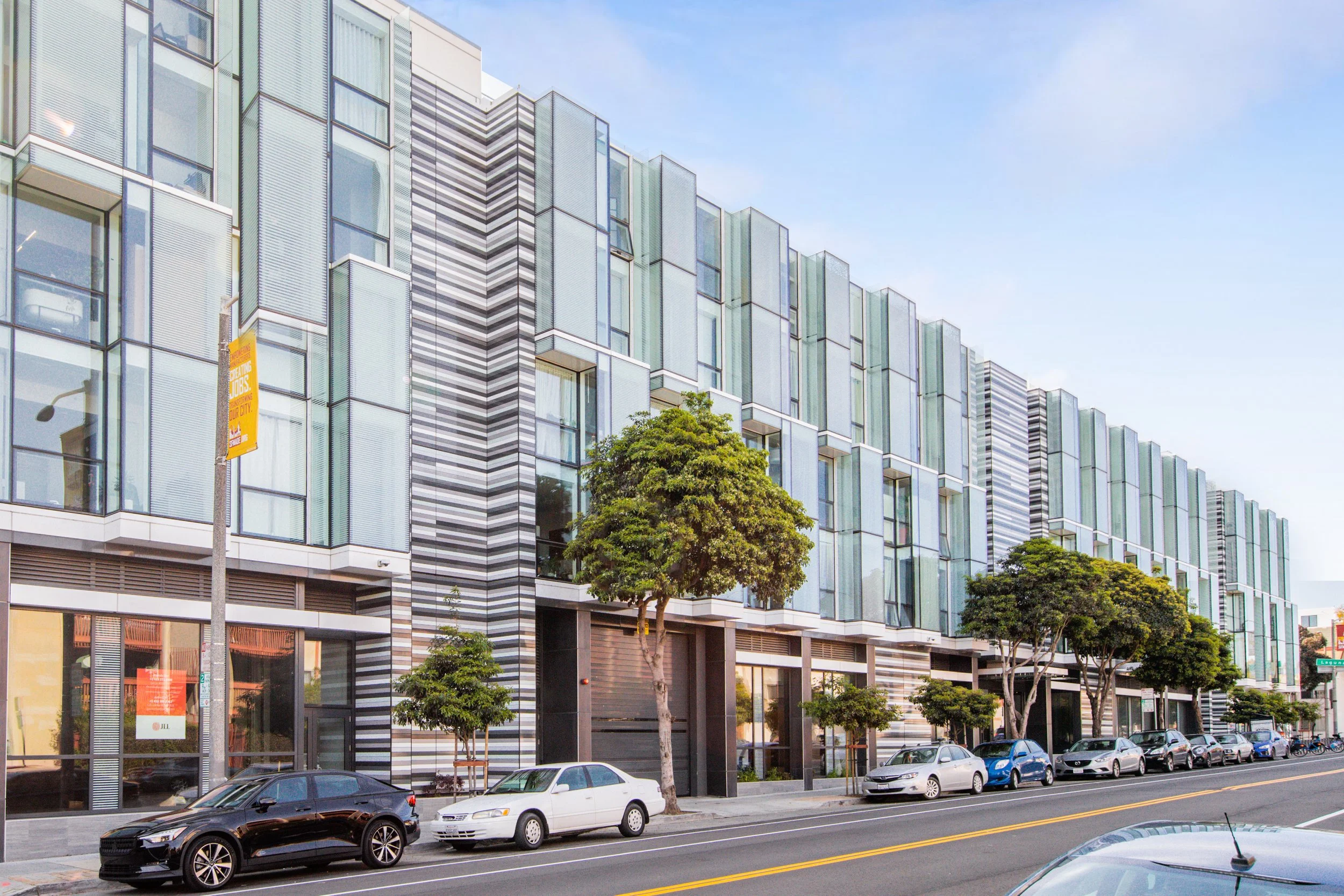 A modern multi-story building with glass windows and striped design, lined with trees on the sidewalk and parked cars along the street.