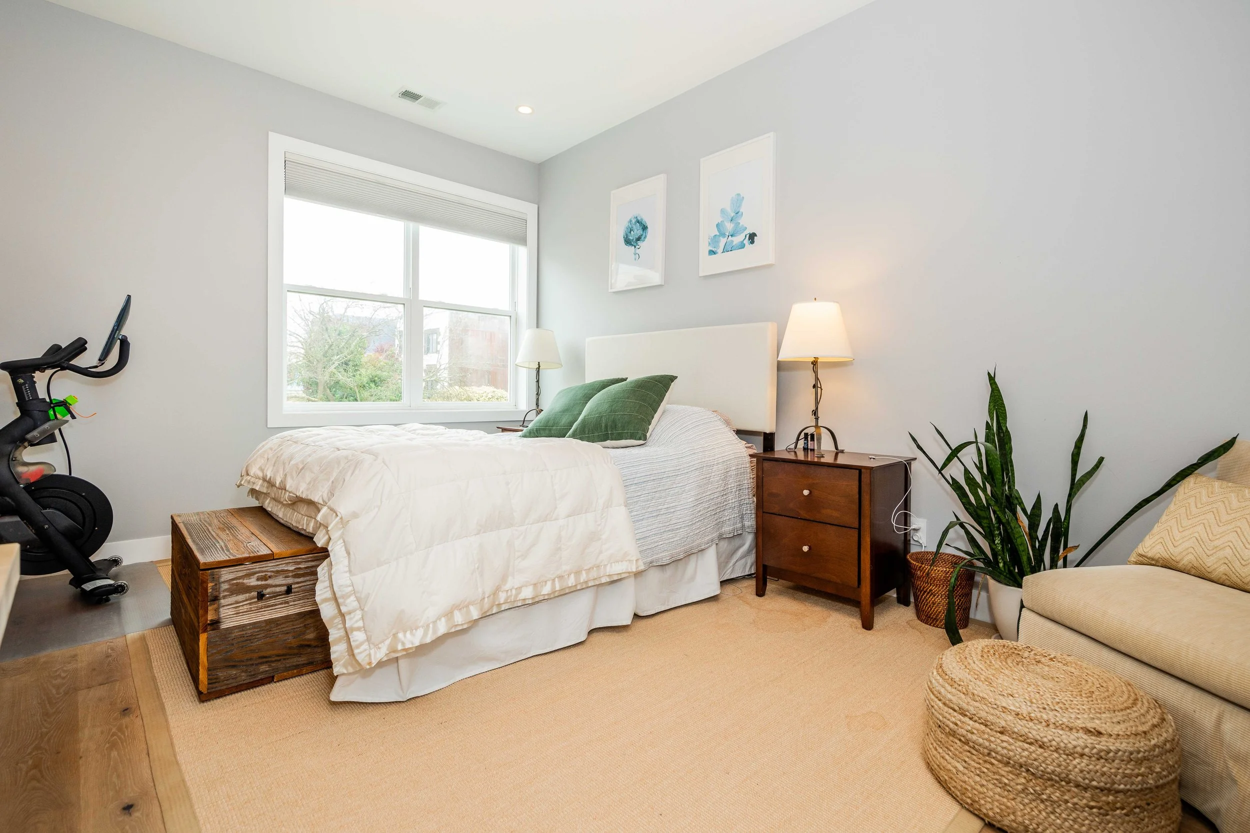 A bedroom with a bed, nightstand, lamps, a potted plant, and a treadmill near the bed.