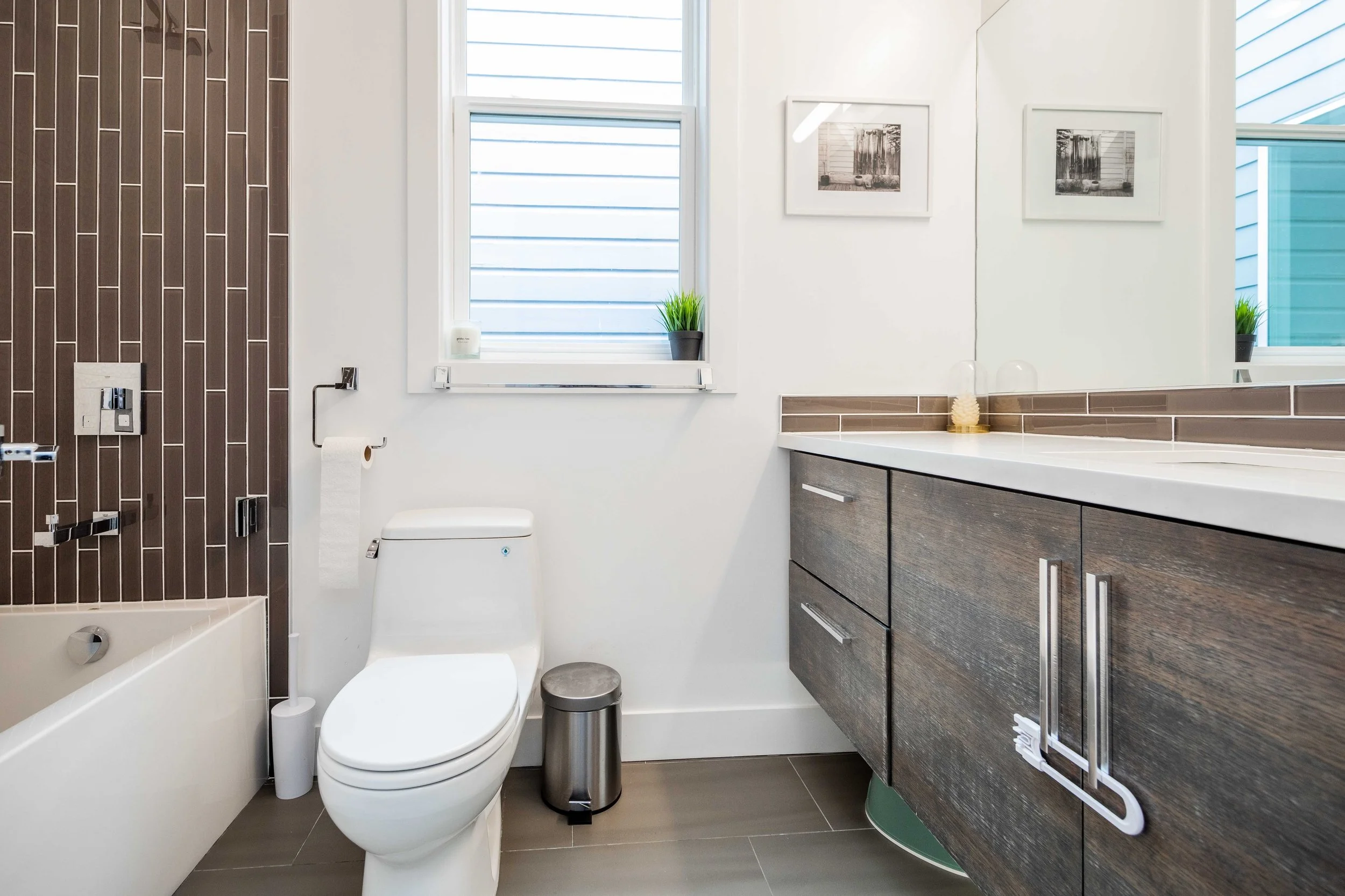 Modern bathroom with a white toilet, a small silver trash can, a wooden vanity with a white countertop, a large mirror, and two framed black-and-white pictures, with window blinds and small potted plants.