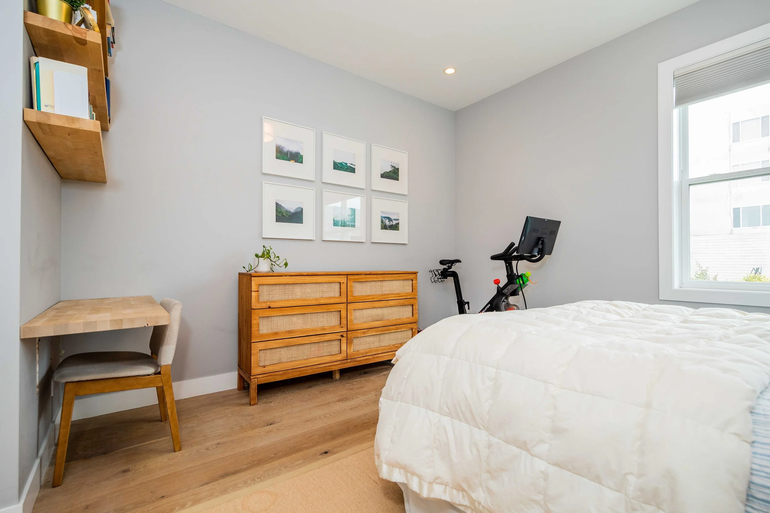 A bedroom with a bed, a wooden dresser, and a stationary exercise bike in front of a window, with framed photos on the wall and a small desk with a chair in the corner.