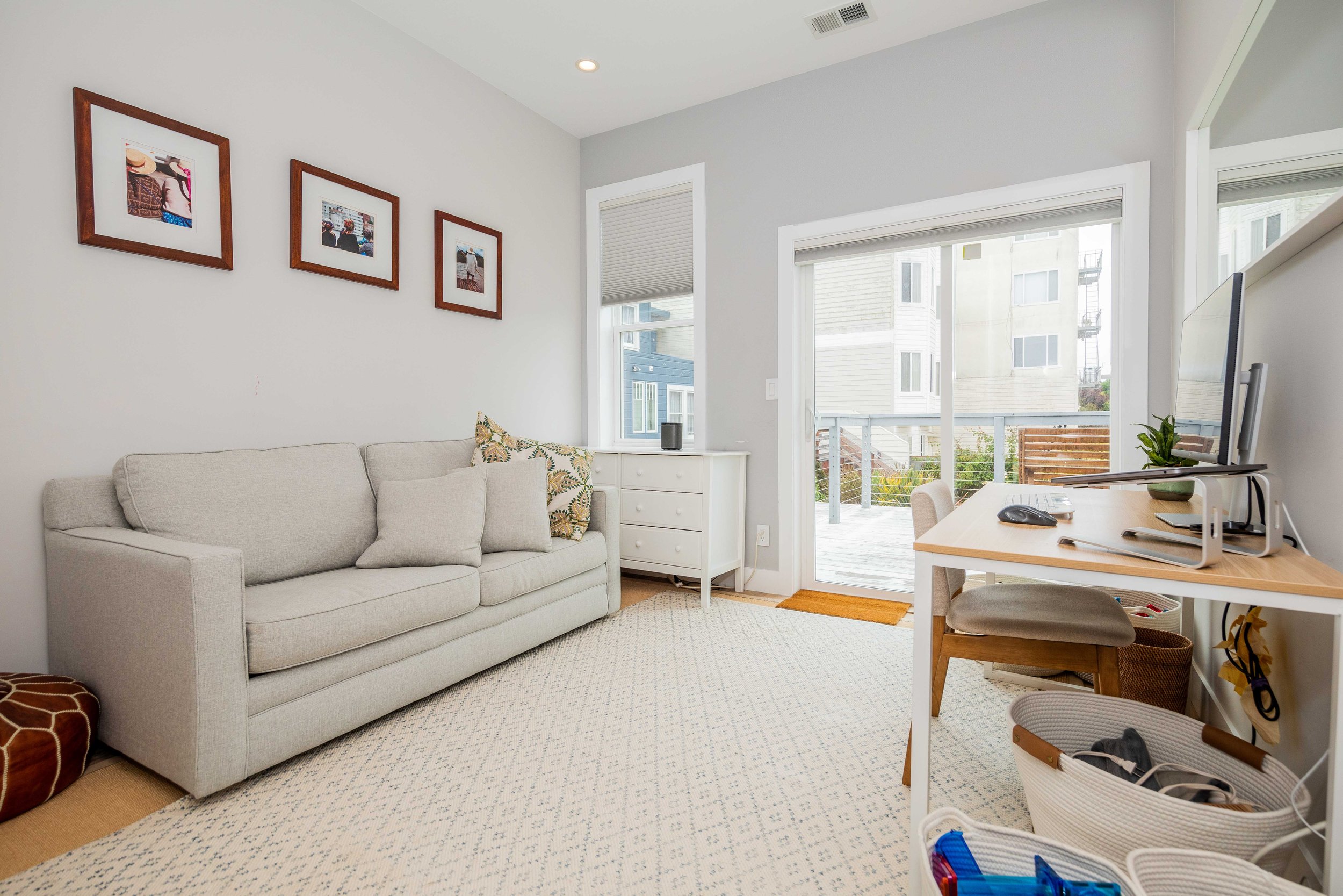 Living room with a white sofa, framed photos on the wall, a sliding glass door leading to a balcony, and a desk with a computer and a chair.