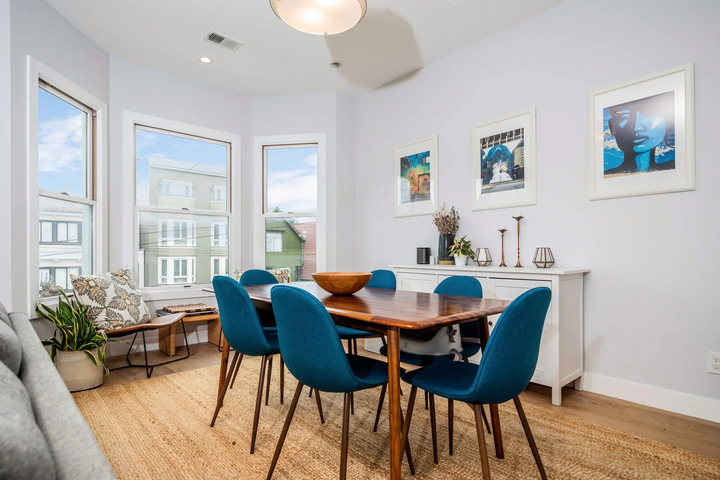 Dining room with a wooden table and six blue upholstered chairs, a white sideboard with decorative items, colorful framed artwork on the white wall, windows showing neighboring houses, a potted plant, and a beige rug.