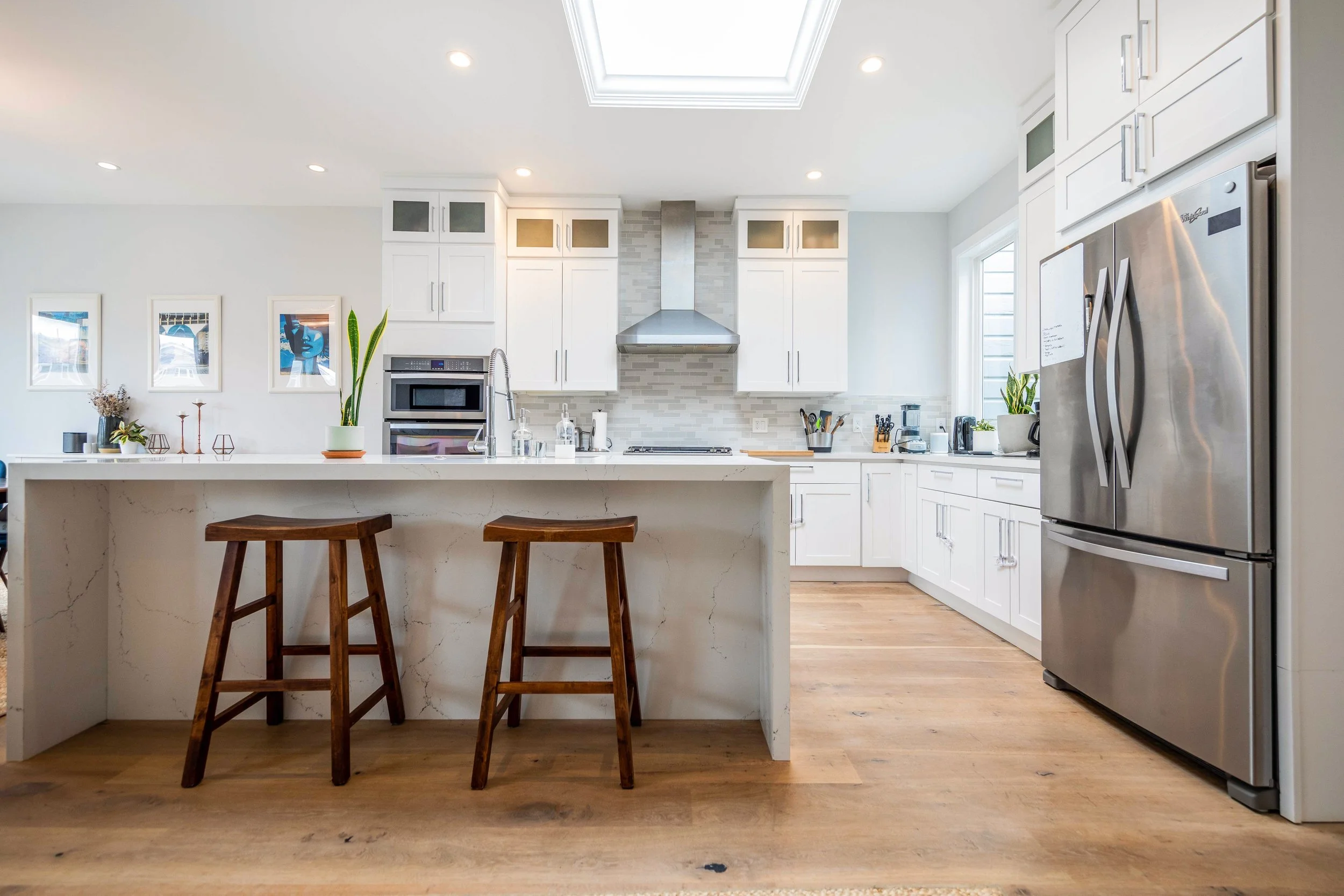 Modern white kitchen with stainless steel appliances, a large island with a marble countertop, and two wooden bar stools. Built-in oven, microwave, and refrigerator are visible, with a skylight overhead.