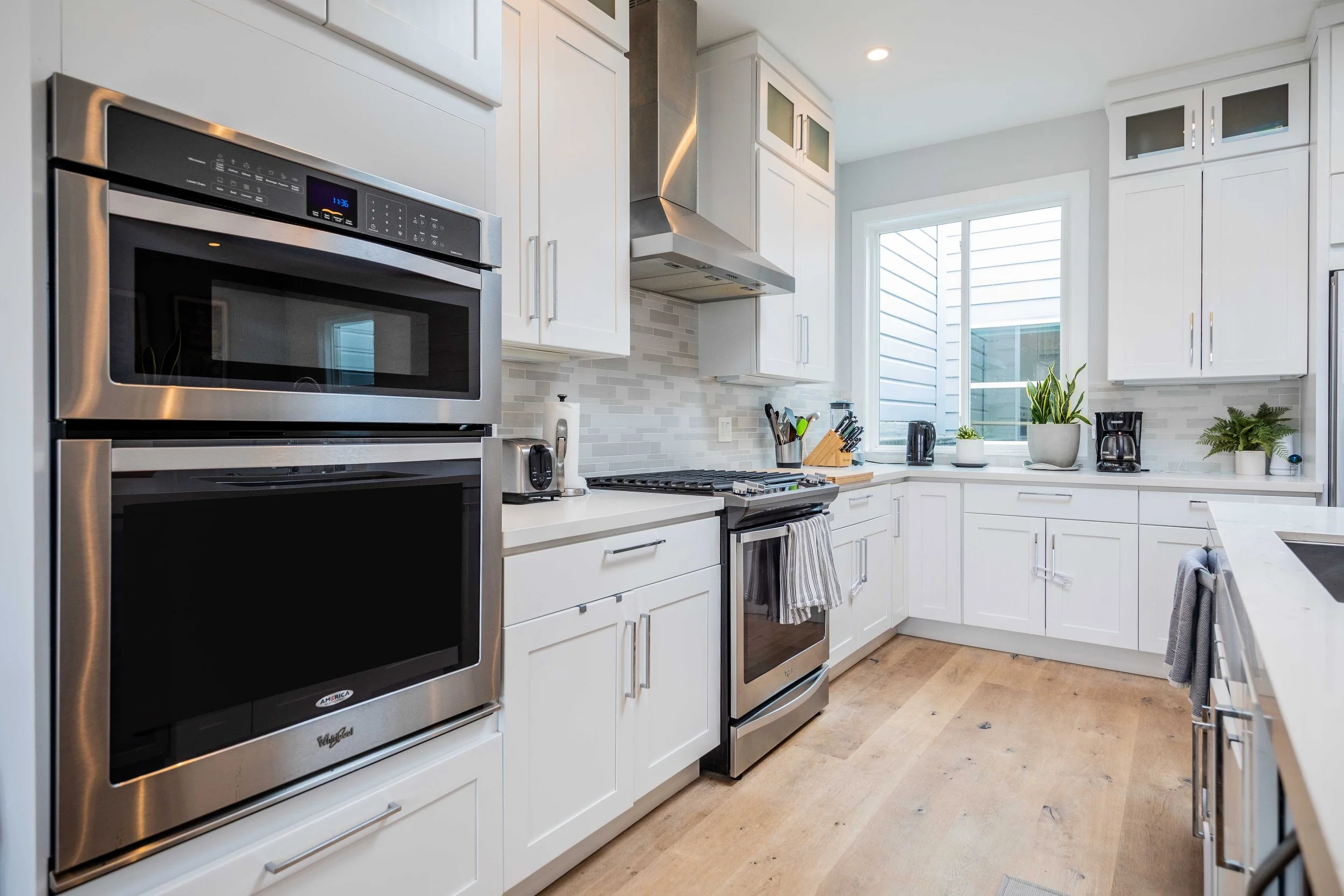 Modern white kitchen with stainless steel appliances, including a microwave, oven, and stove. There are white cabinets, a window with sunlight, and various potted plants and kitchen tools on the counters.