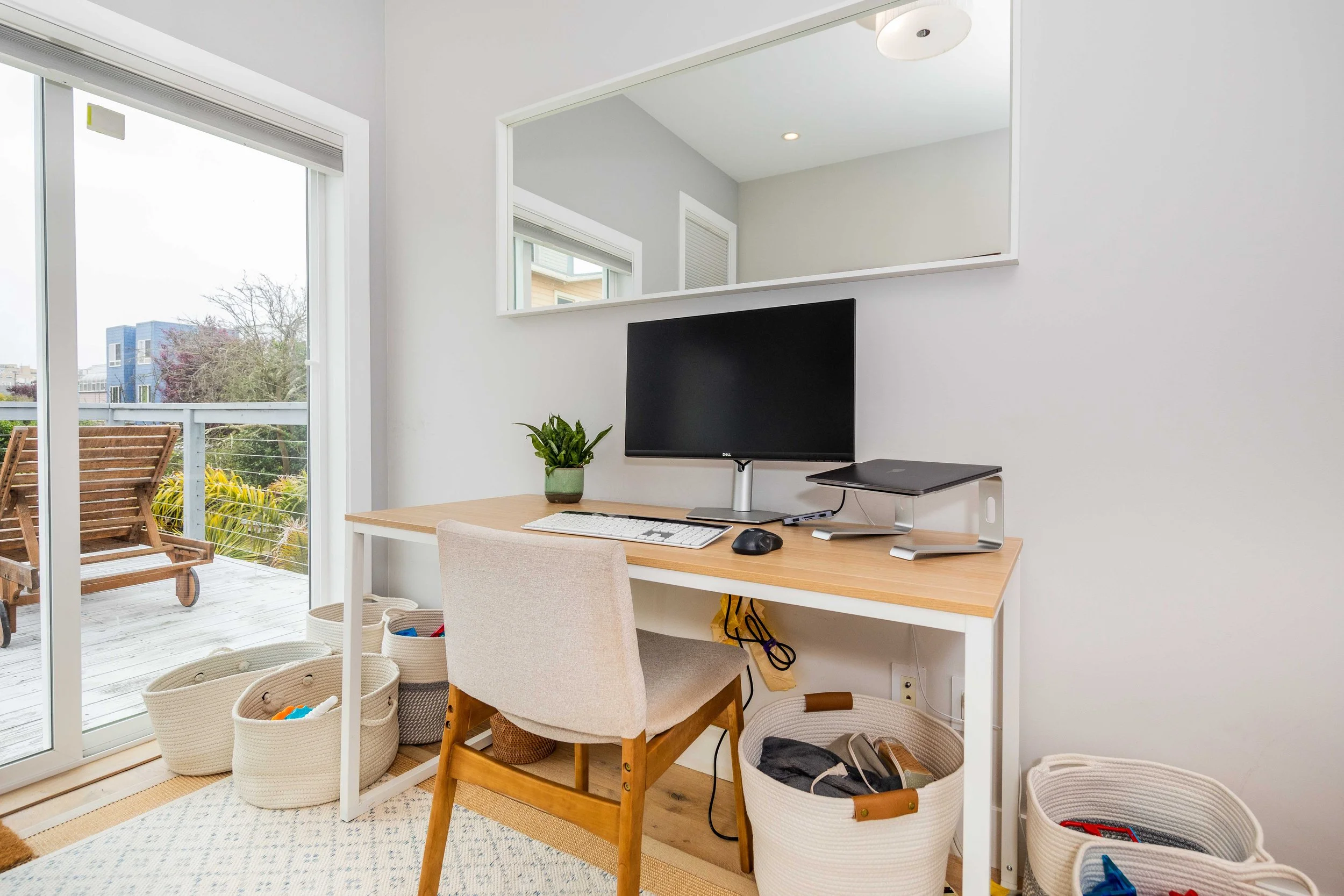 Home office setup with a wooden desk, computer monitor, keyboard, mouse, plant, and a gray chair inside a room with a sliding glass door leading to an outdoor balcony with wooden chairs and greenery.