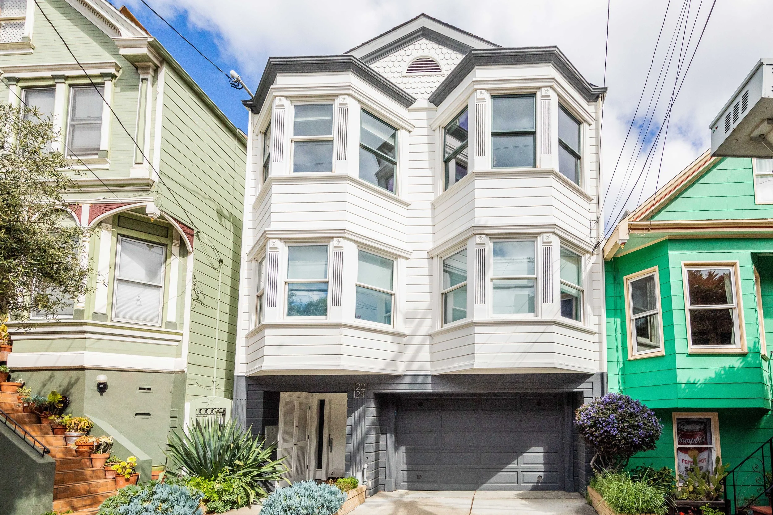 A modern white multi-story house with bay windows, situated between two colorful houses, one green and one turquoise, featuring a garage and a small garden with plants and flowers in the front.