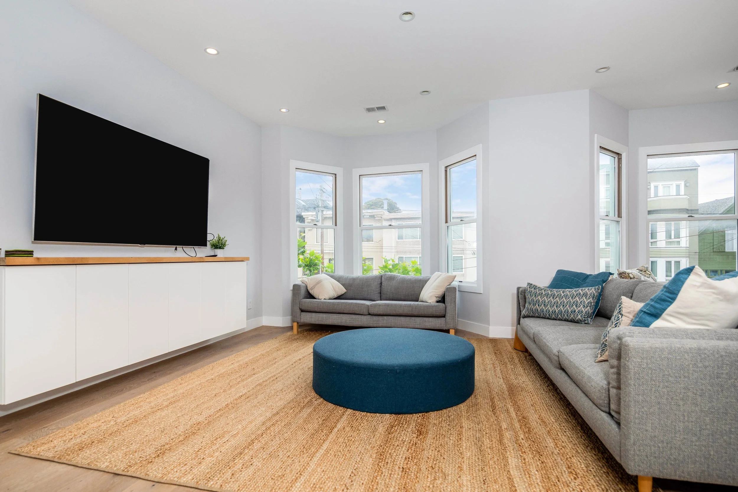 A modern living room with a wall-mounted flat-screen TV, gray sofas with blue and patterned pillows, a round blue ottoman, a beige natural fiber rug, and multiple windows with views of neighboring buildings and greenery.