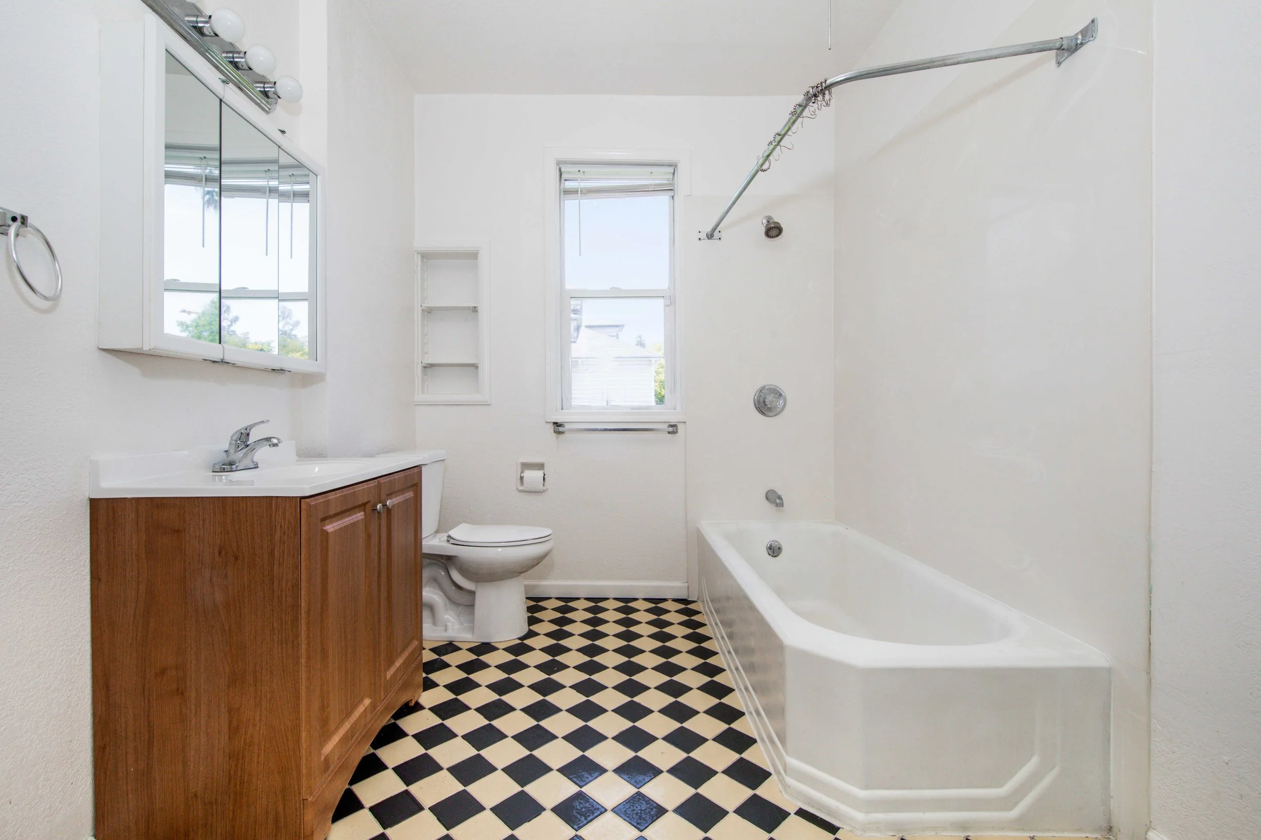 A small bathroom with a wood vanity, a toilet, a bathtub, and two windows. The floor has black and white checkered tiles.