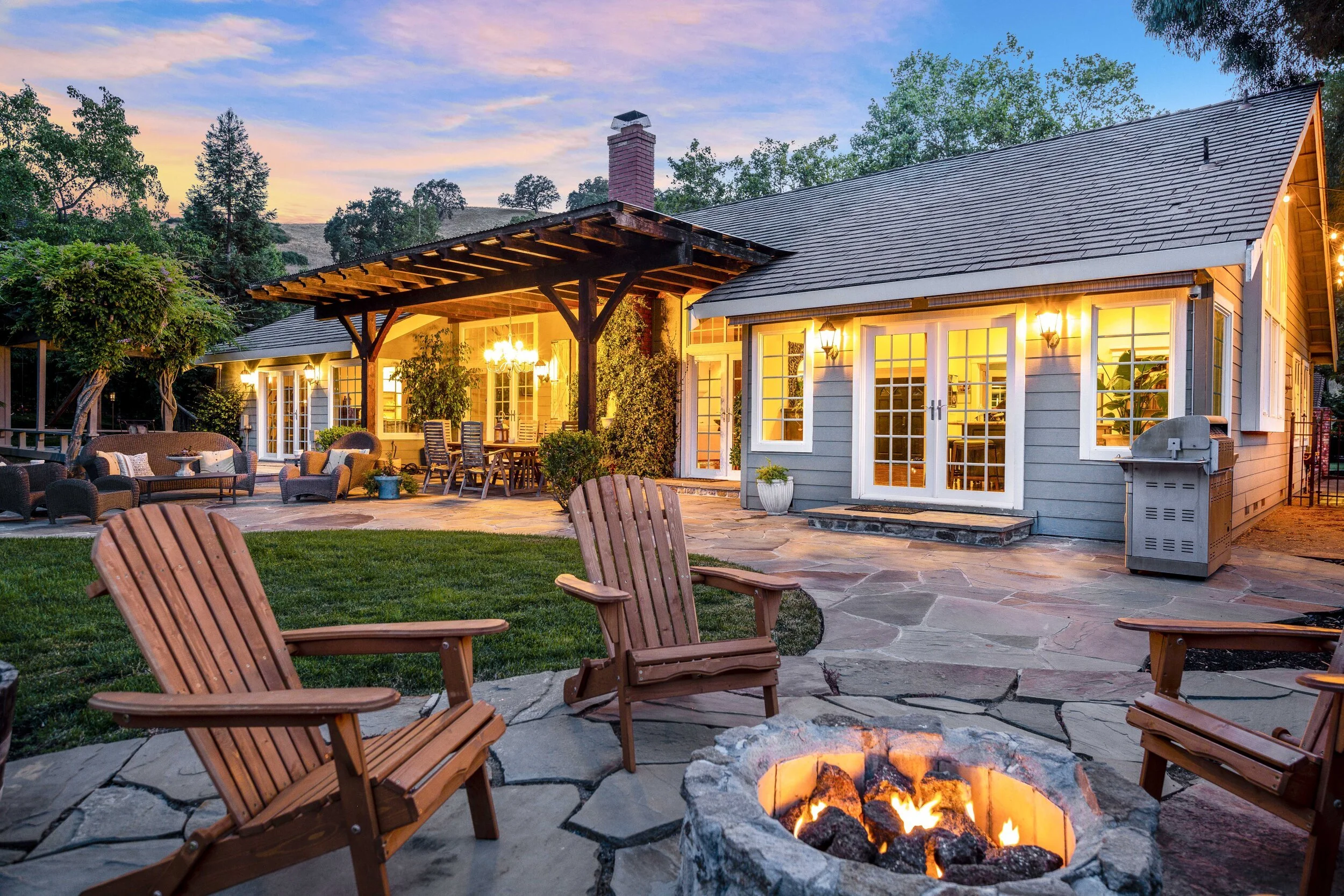 A backyard patio at dusk with a fire pit, Adirondack chairs, outdoor furniture, and a house with lit windows and French doors.