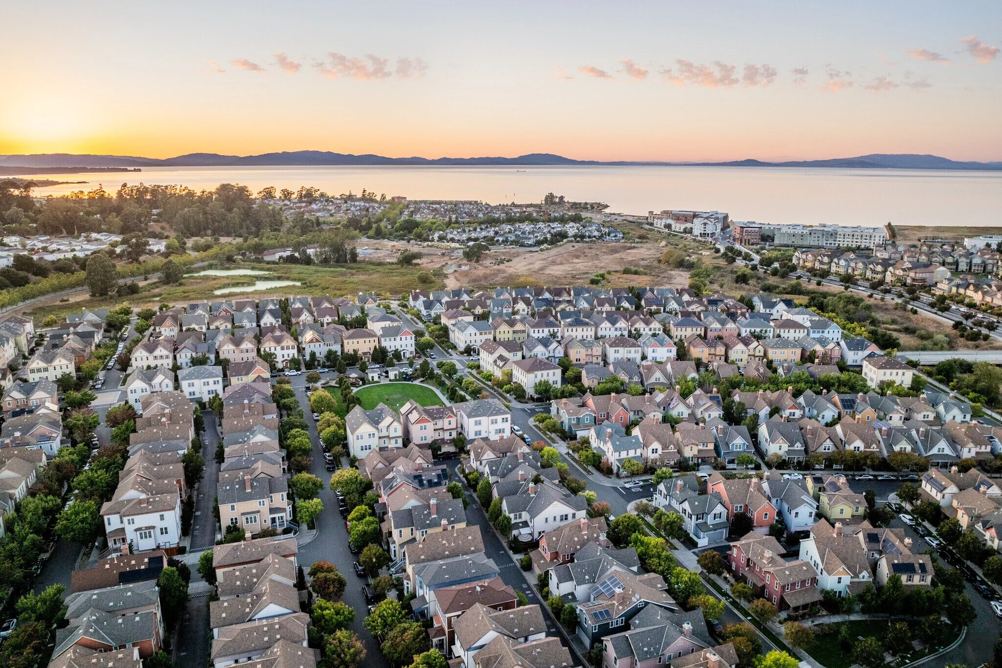 Aerial view of a residential neighborhood near a large body of water at sunset, with houses, trees, and streets.
