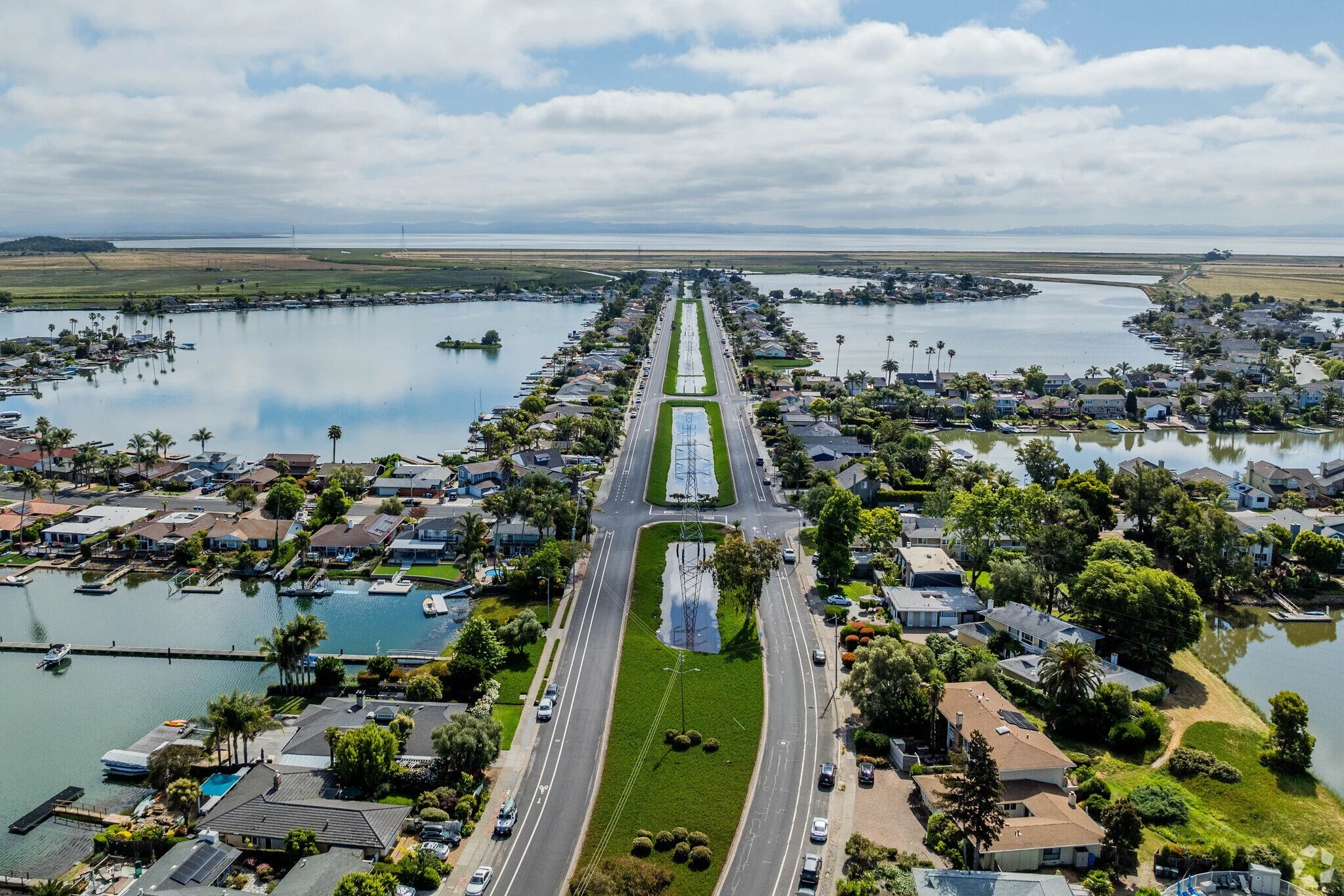 An aerial view of a coastal residential neighborhood with houses, water canals, and pilings, featuring a road with landscaped medians and reflecting pools running through the center, under partly cloudy skies.