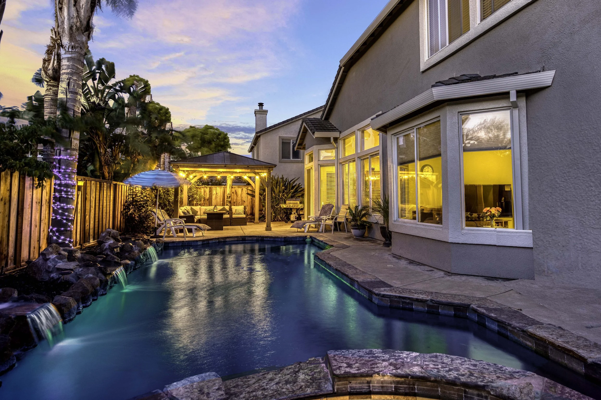 A backyard scene during sunset featuring a swimming pool with waterfalls, a wooden deck with outdoor furniture and string lights, a gazebo, and a house with large windows glowing with warm interior light, surrounded by trees and a wooden fence.