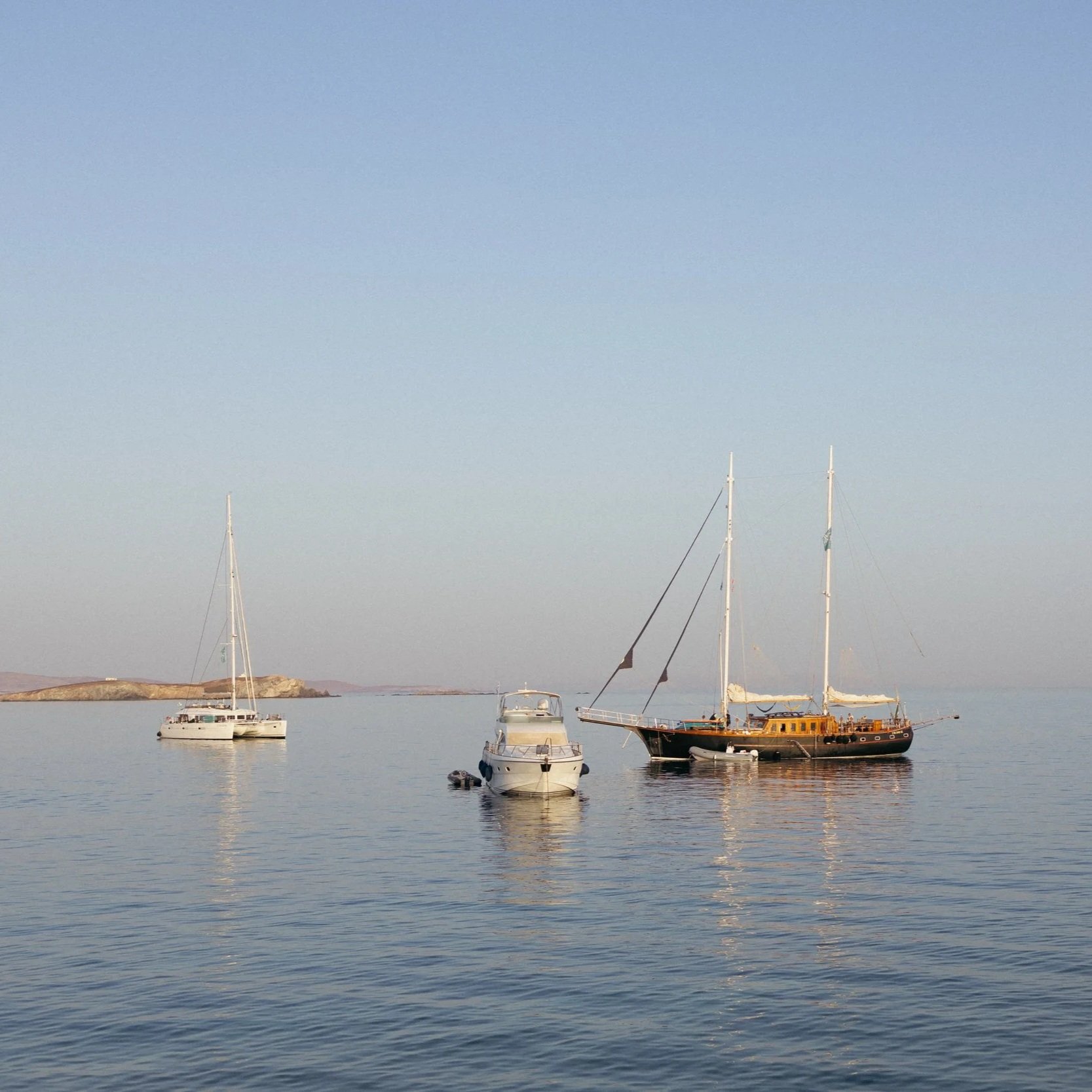 Three boats floating on calm water near a rocky shoreline with a small island in the background and a clear sky above.