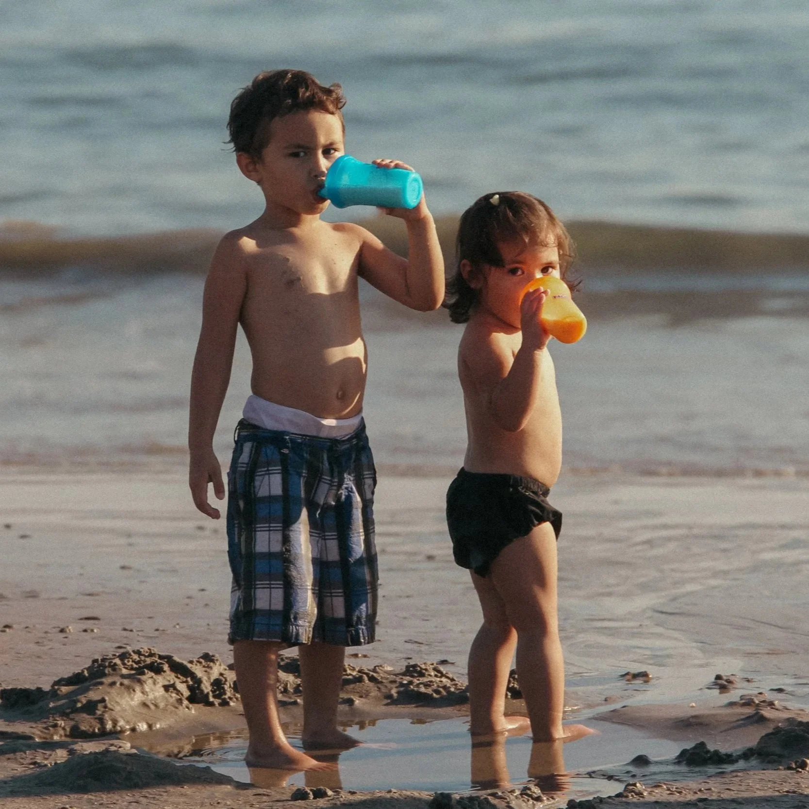 Two young children, a boy and a girl, standing on the beach near the water, drinking from plastic bottles.