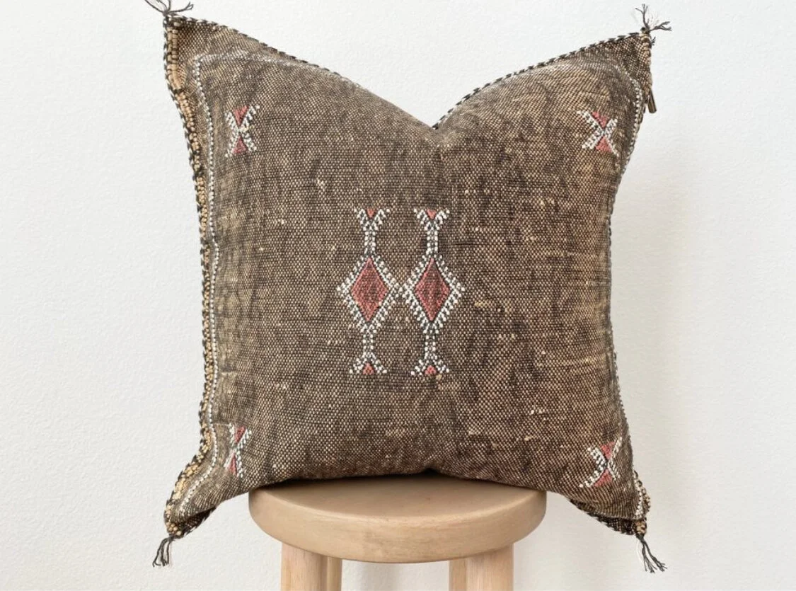 Moroccan silk pillow in brown sitting on stool on white background.