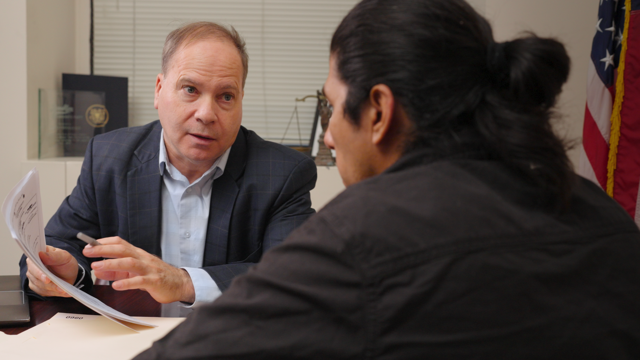 A man in a suit consulting with another man with long black hair in an office.