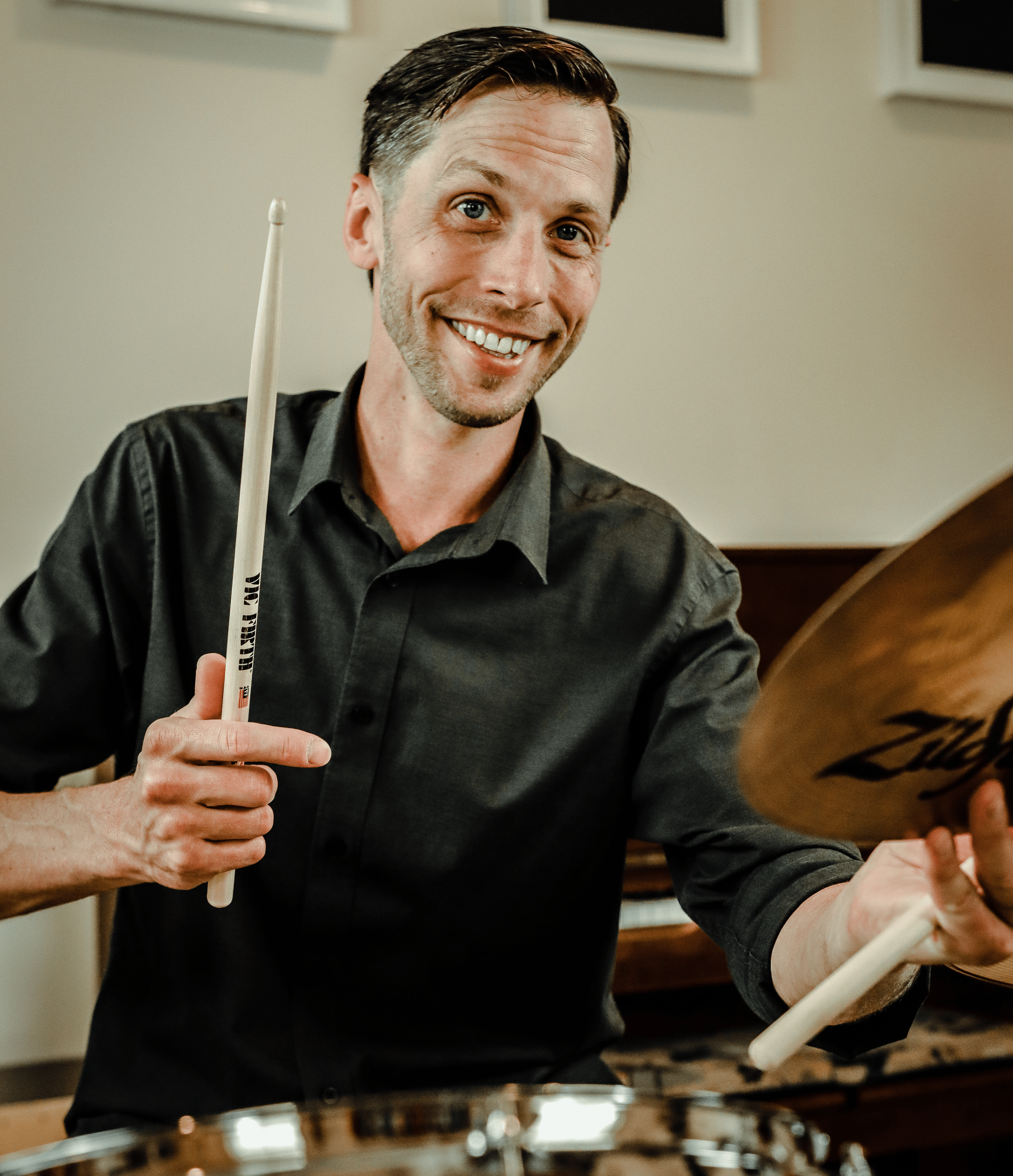Phil Didlake smiling holding drum sticks, cymbals with a drum set a piano in the background
