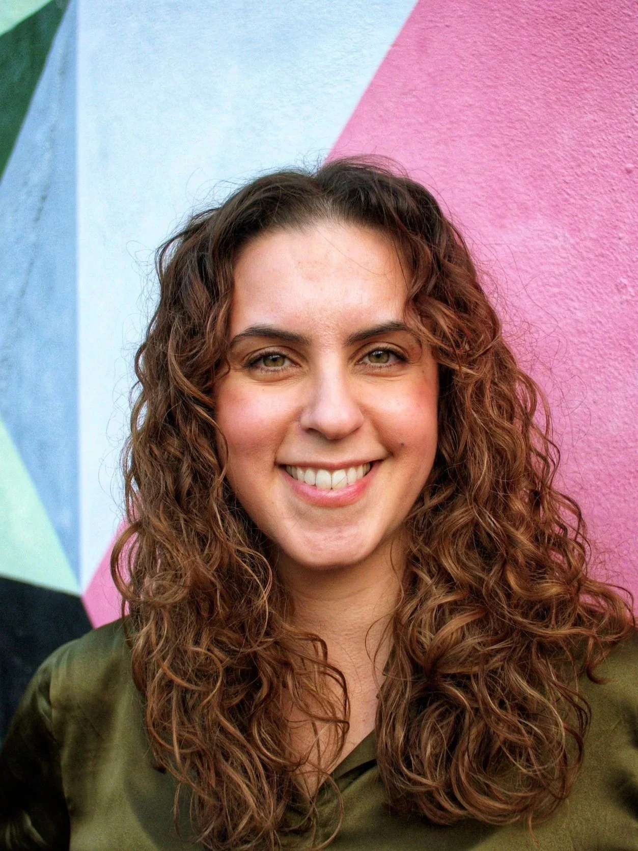 A woman with curly brown hair smiling in front of a colorful geometric mural wall.