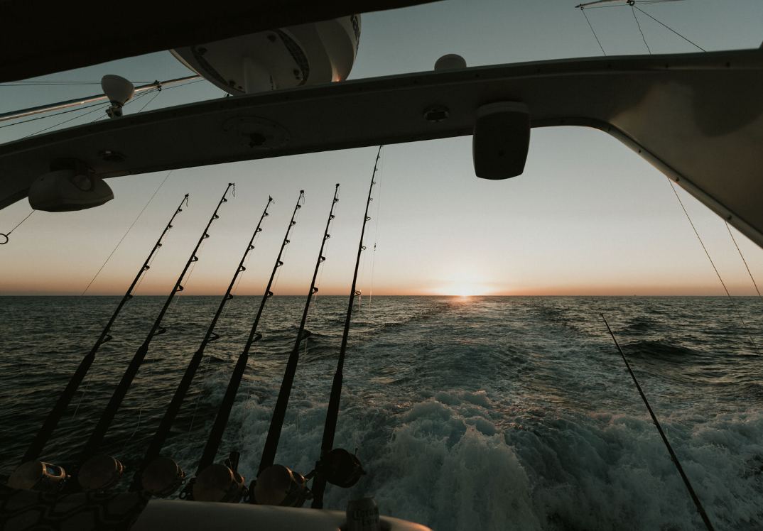 Vista del mar desde la parte trasera de un barco de pesca, con cañas de pescar en soporte y el sol en el horizonte al atardecer.