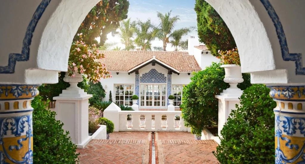 Vista de una casa estilo mediterráneo, con entrada mediante un arco decorado, jardines y plantas verdes, y un camino de ladrillo.