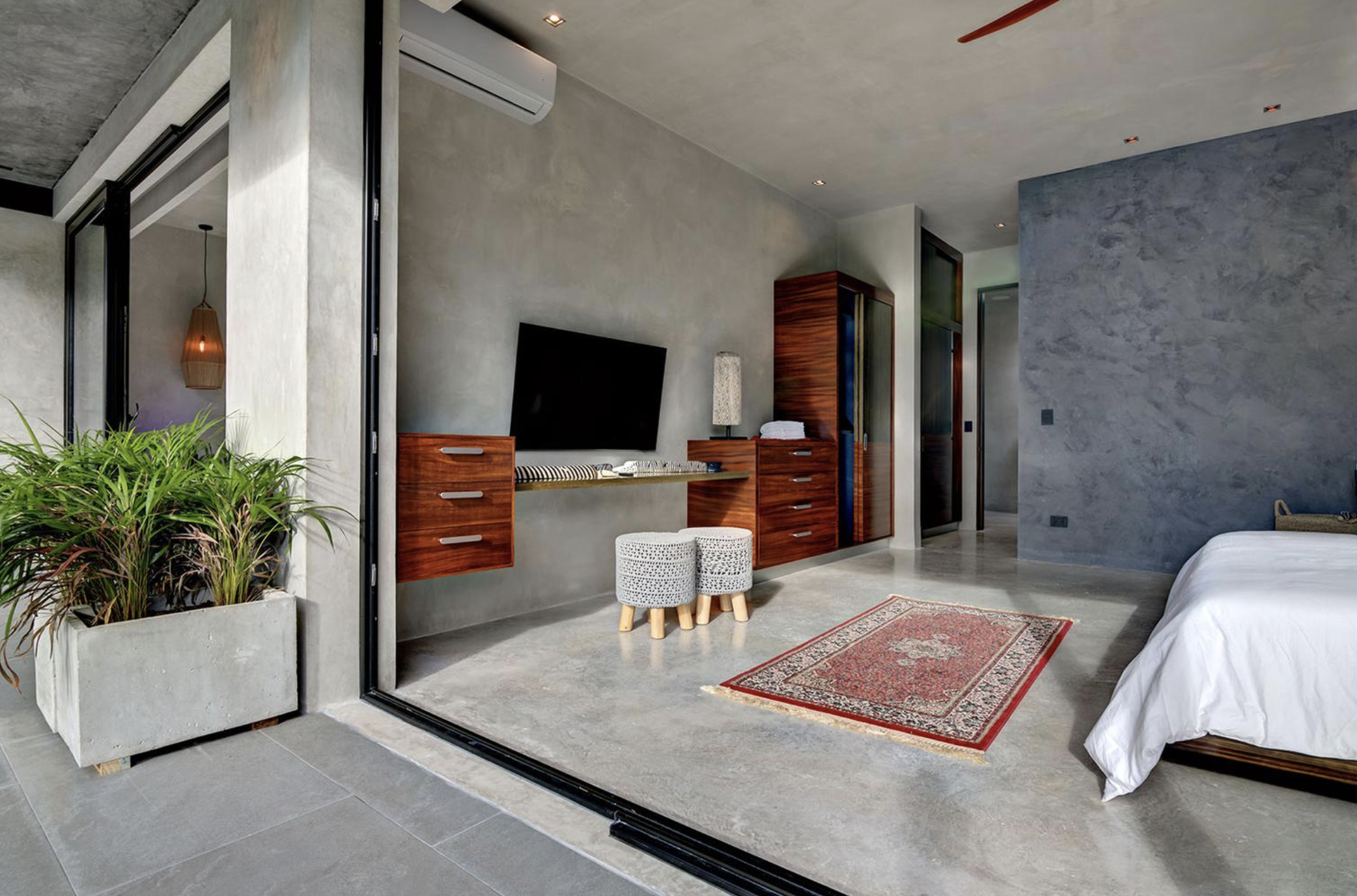 Habitación moderna con paredes de concreto, piso de cemento, una cama con sábanas blancas, un área de estar con TV y muebles de madera, y plantas en una maceta de concreto.