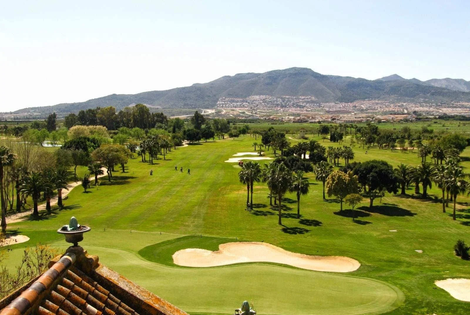Vista panorámica de un campo de golf con árboles, hoyos y arena, rodeado de montañas al fondo bajo un cielo despejado.