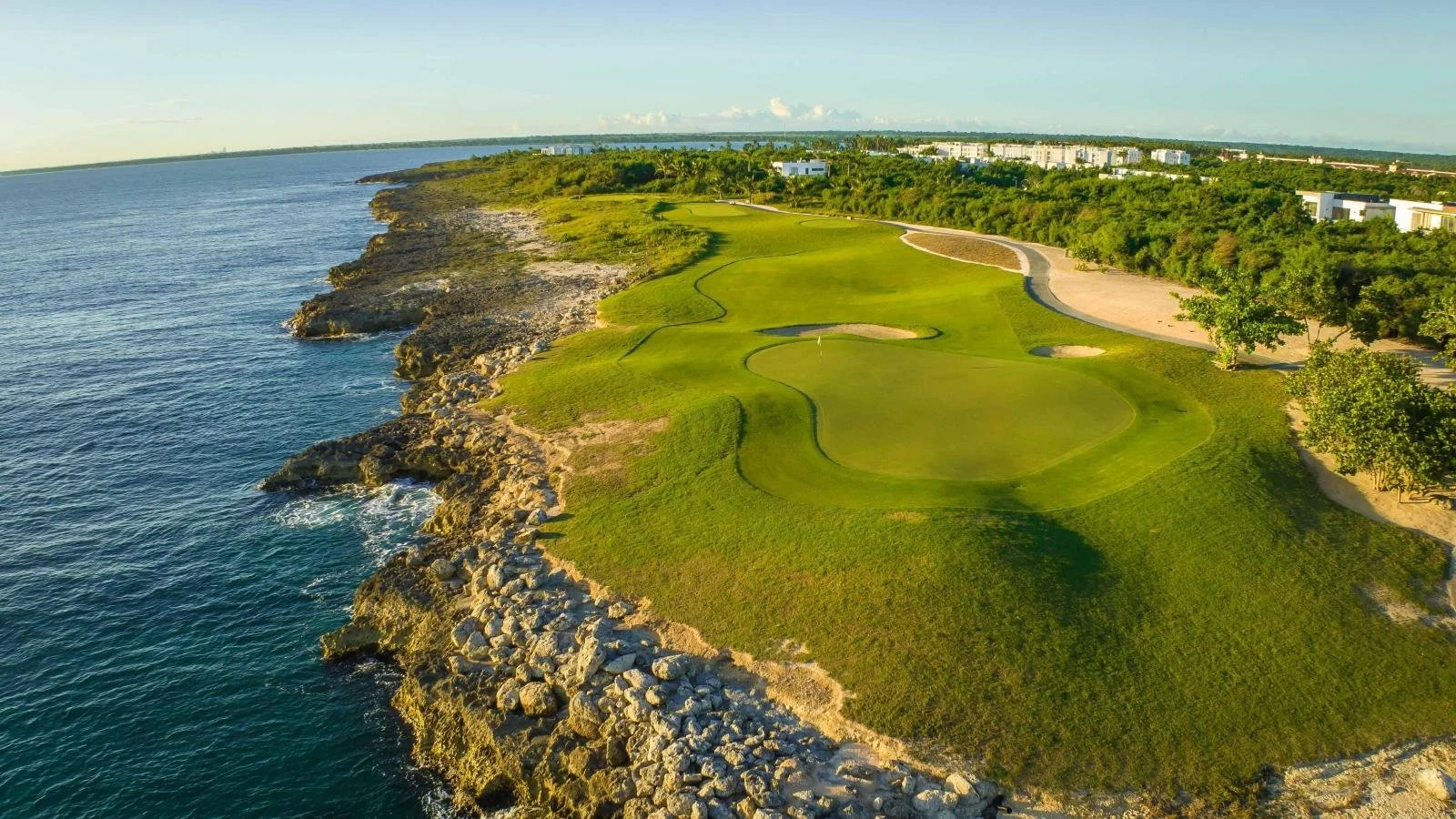 Campo de golf junto al mar con césped verde y rocas en la costa, árboles y edificios en el fondo con cielo despejado.