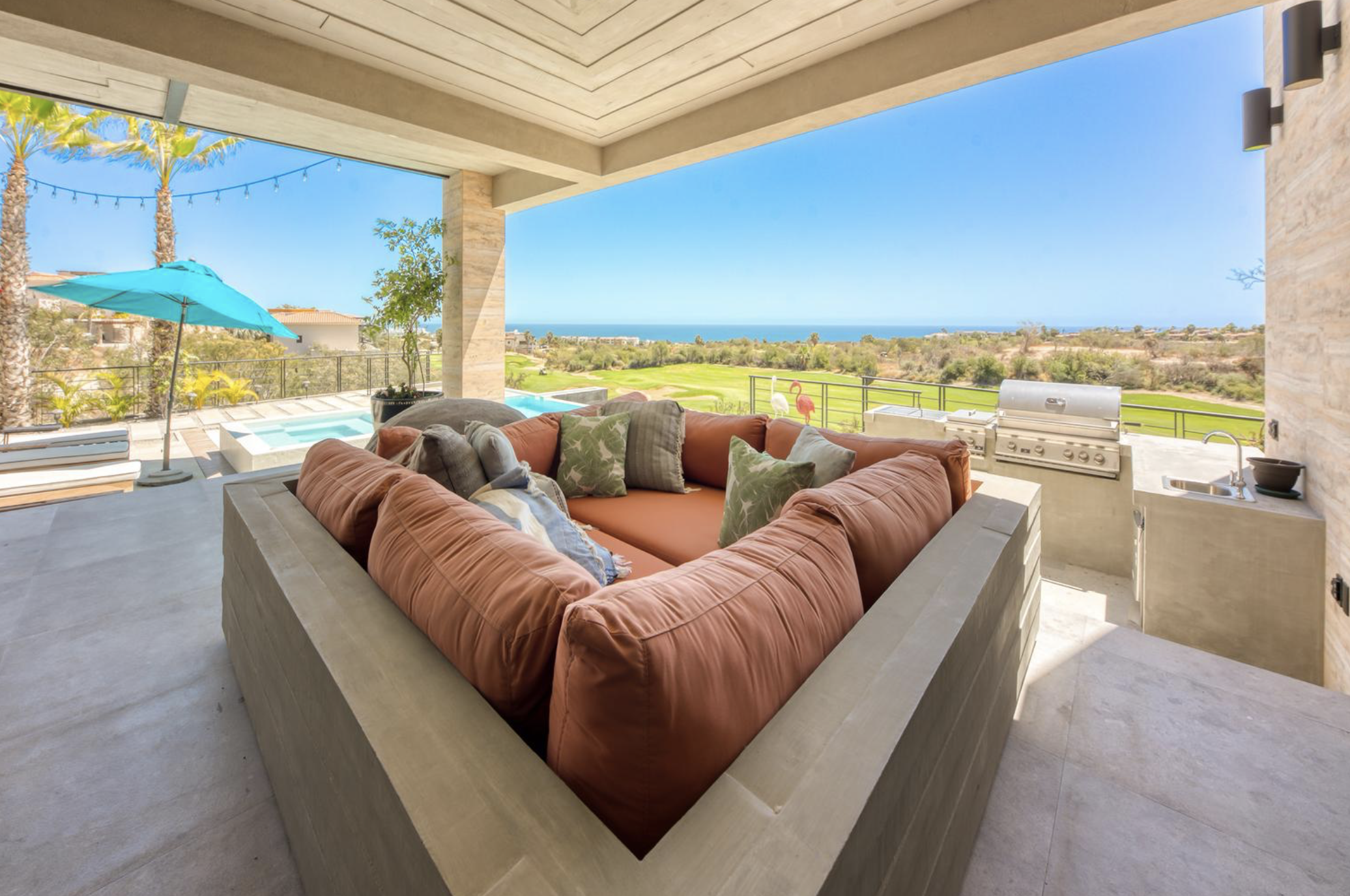 Espacio al aire libre con vista al mar, área de descanso con sofá en forma de L con cojines coloridos, sombrilla azul, área de barbacoa y piscina pequeña en el fondo.