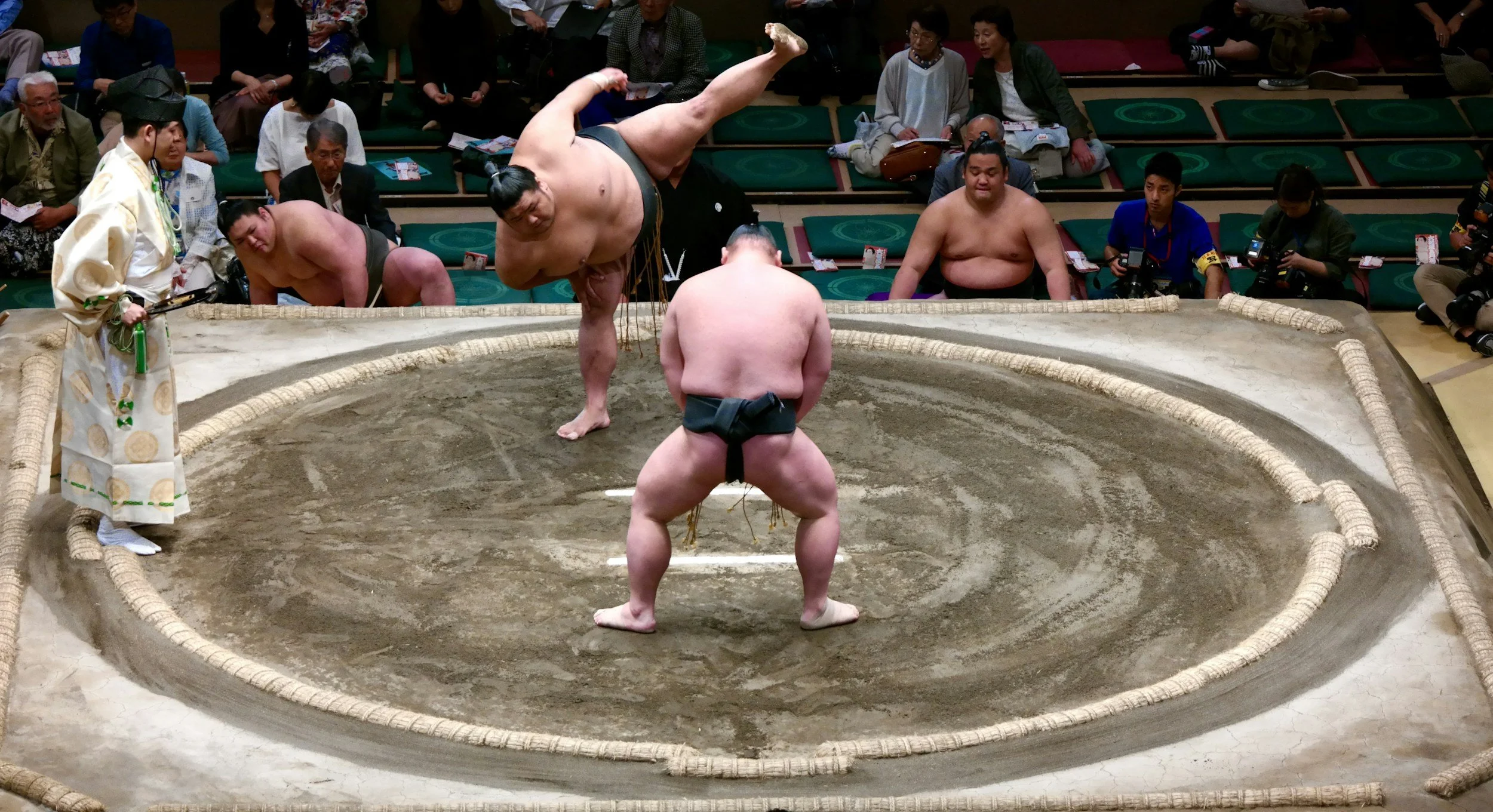 Dos luchadores de sumo compitiendo en un ring tradicional, con un árbitro observando y espectadores en el fondo.