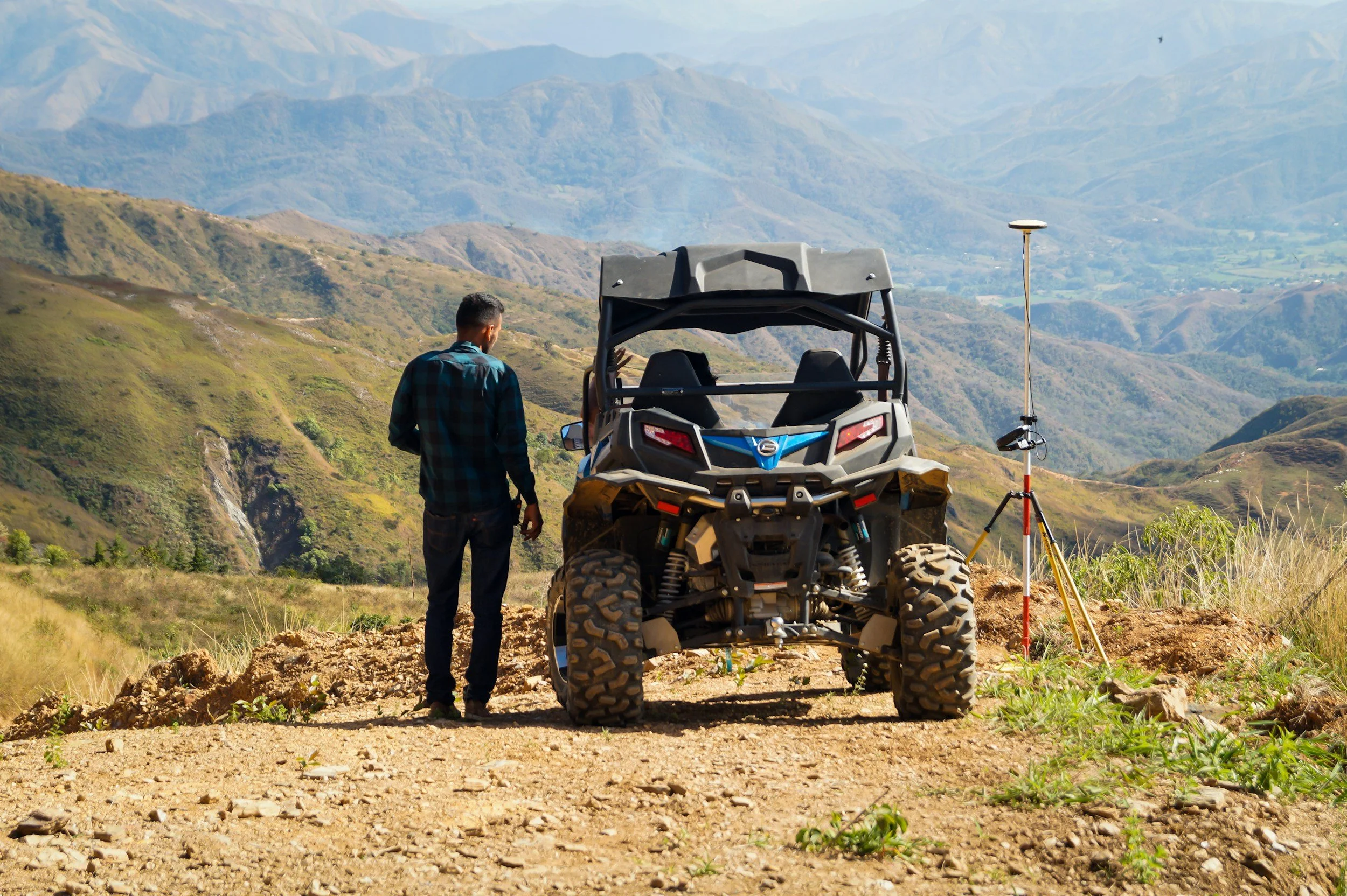 Un hombre de espaldas mirando un vehículo todo terreno en un paisaje montañoso con un equipo de medición en el suelo.