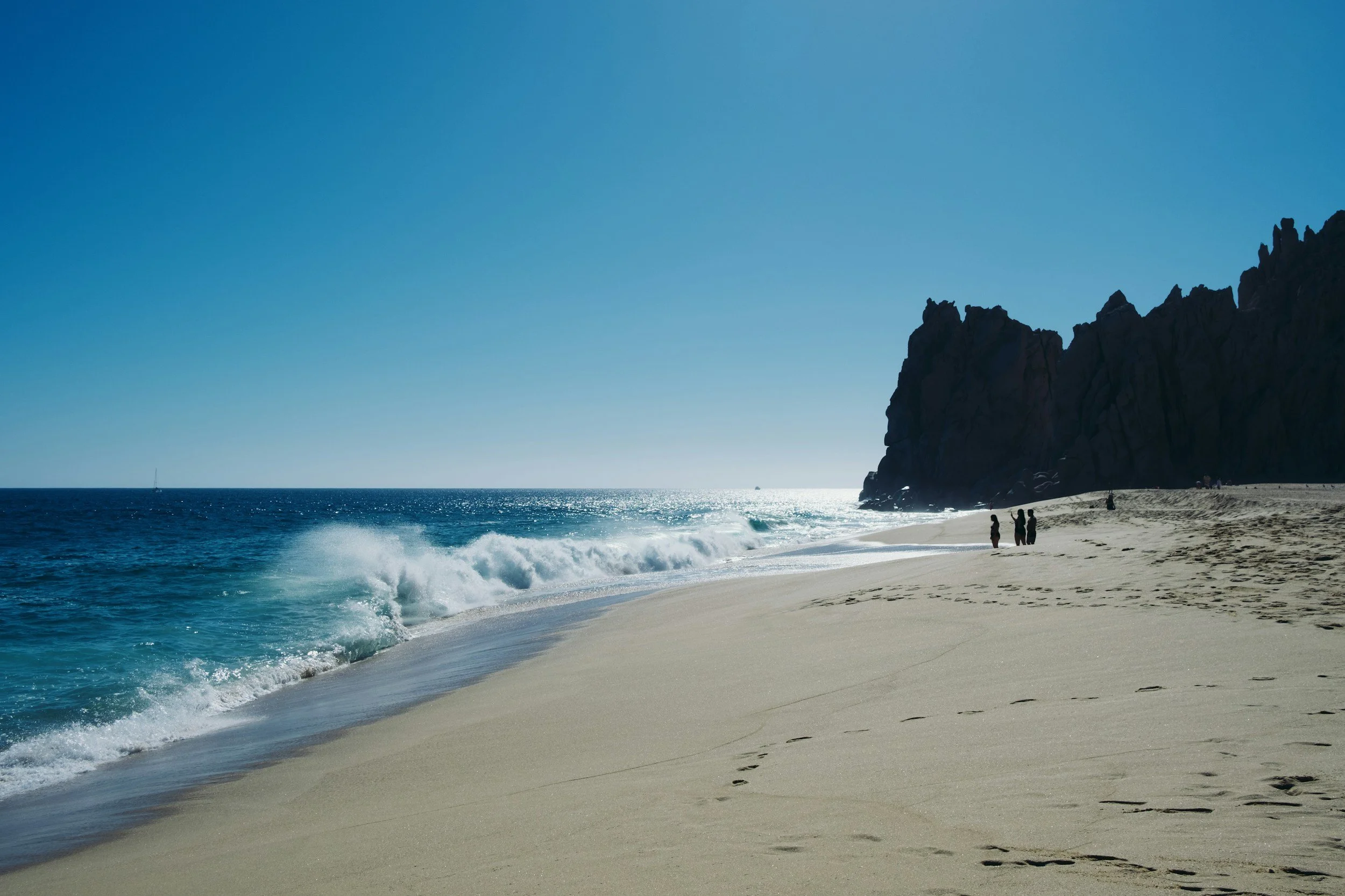 Playa con arena dorada, olas del mar, acantilados oscuros a la derecha y cielo despejado.